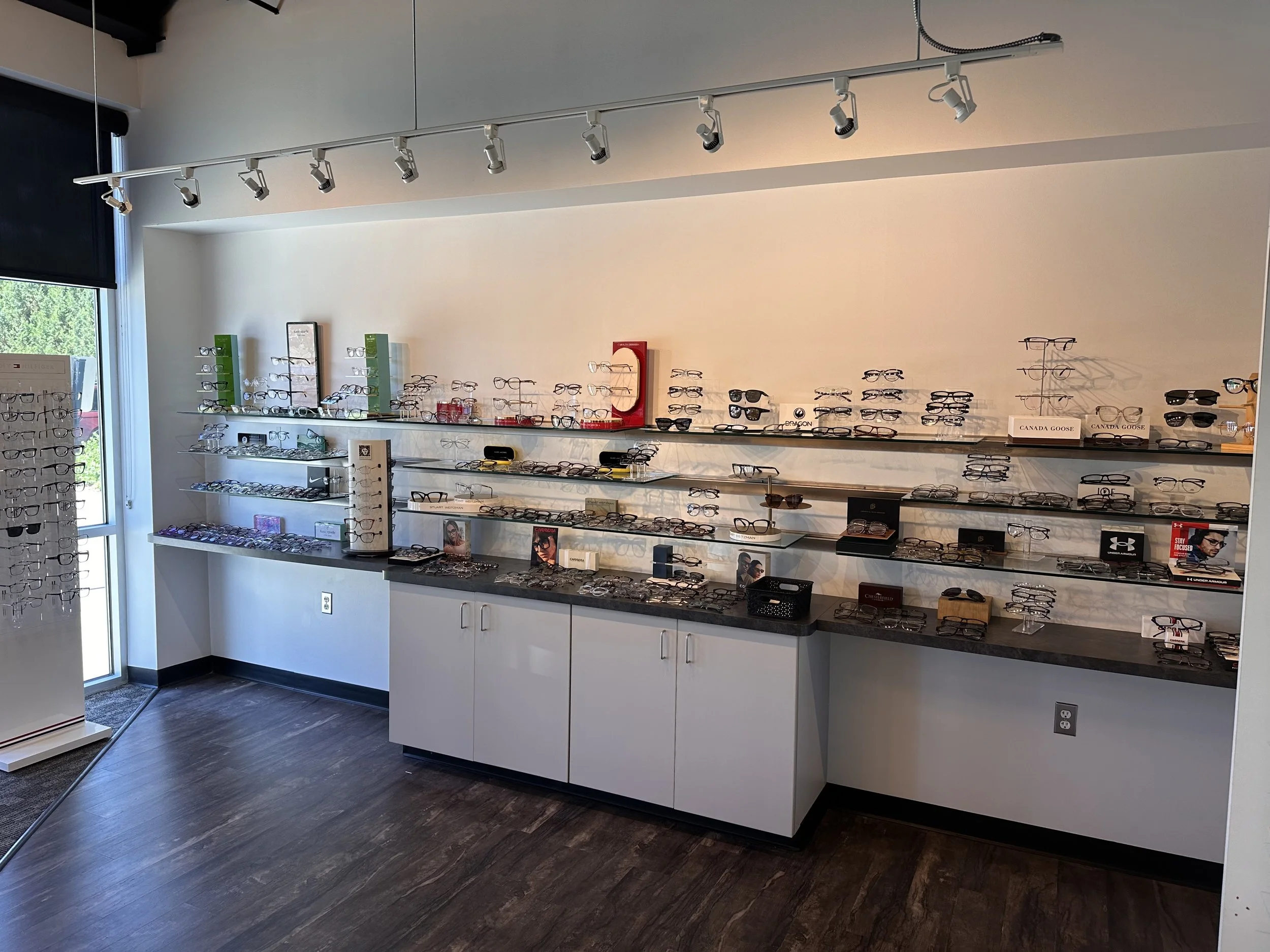 Inside an eyewear store with multiple shelves displaying eyeglasses and sunglasses, a window to the left, and track lighting on the ceiling.