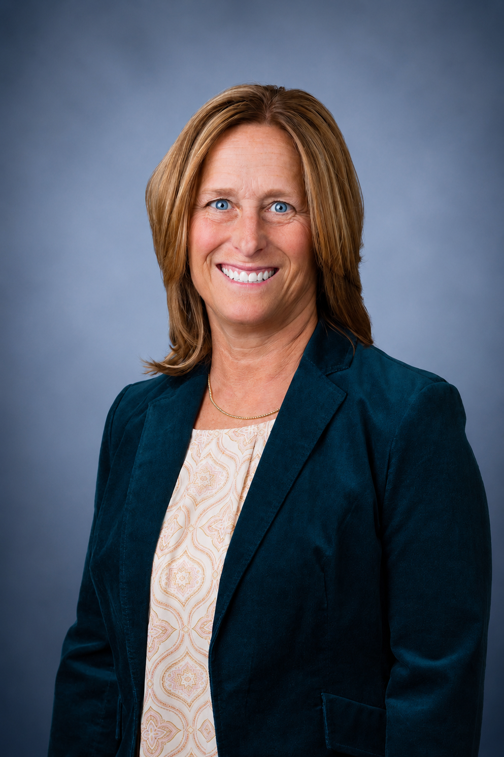 Headshot of a woman with shoulder-length brown hair and blue eyes, smiling, wearing a dark blazer over a patterned blouse, against a gray background.