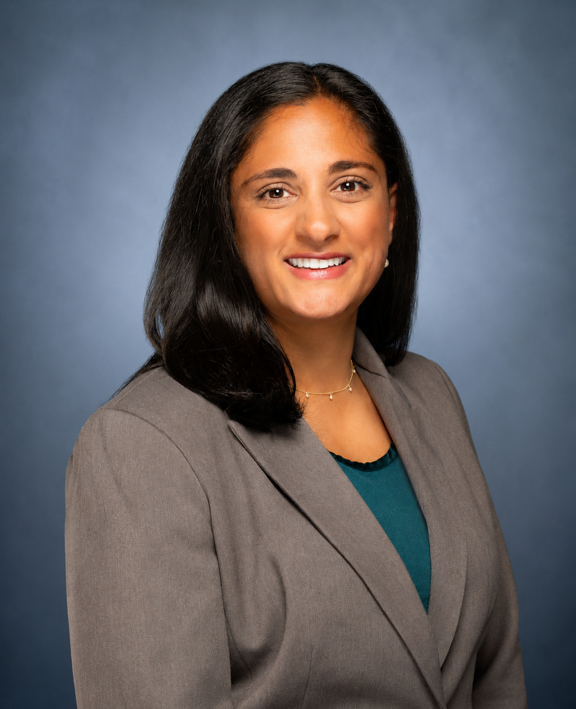 A professional woman with shoulder-length black hair, wearing a gray blazer, blue top, and a delicate necklace, smiling against a blue background.