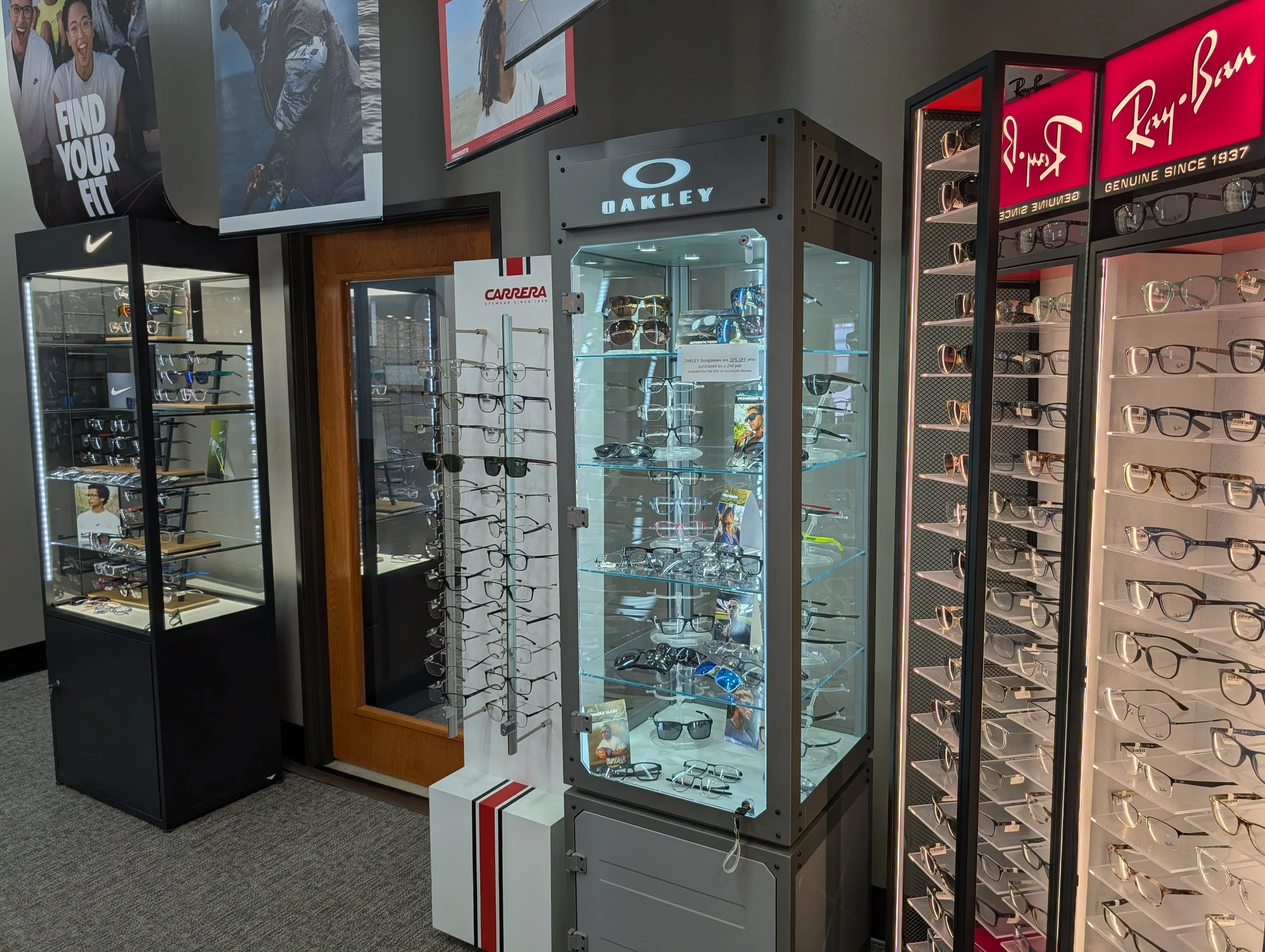 Display of various sunglasses and eyeglasses in retail store with brand signs including Oakley, Carrera, Ray-Ban, and Nike.