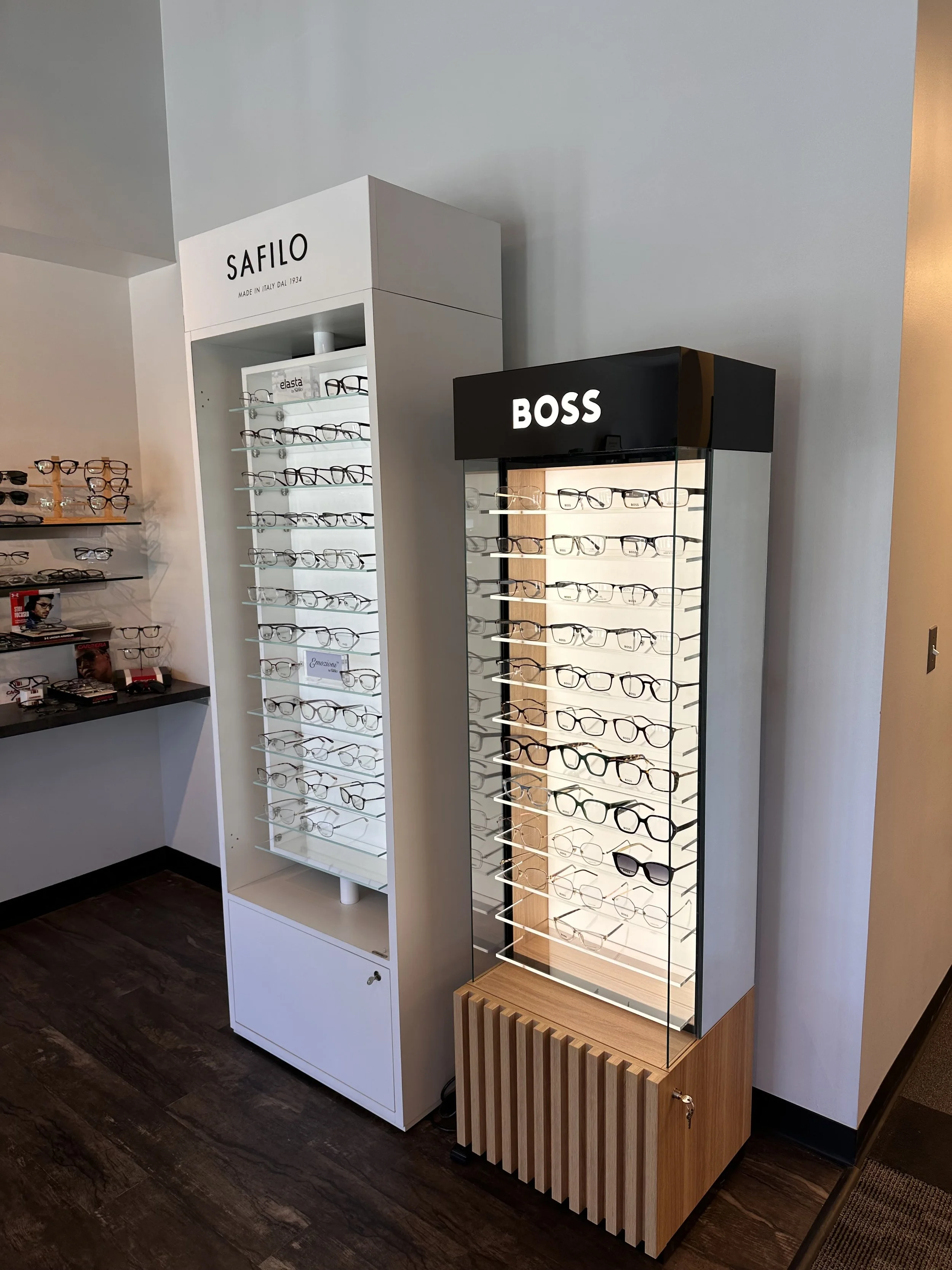 Display of eyeglasses on glass and wood shelves in an optical store with branding signs for Safilo and Boss.