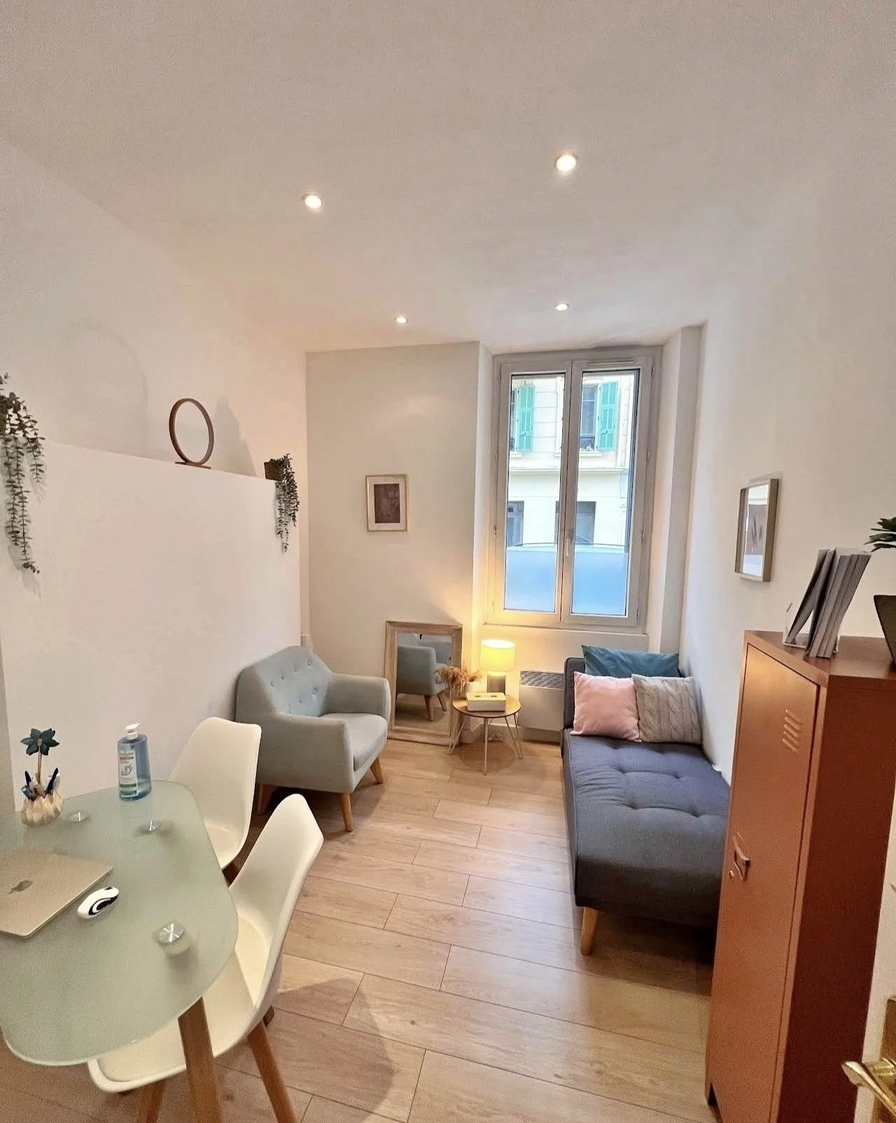 Salon lumineux avec canapé gris, fauteuil beige, table d'appoint avec lampe, fenêtre avec volets bleus, décoration minimaliste, plantes suspendues, table à manger avec chaises blanches, armoire en bois