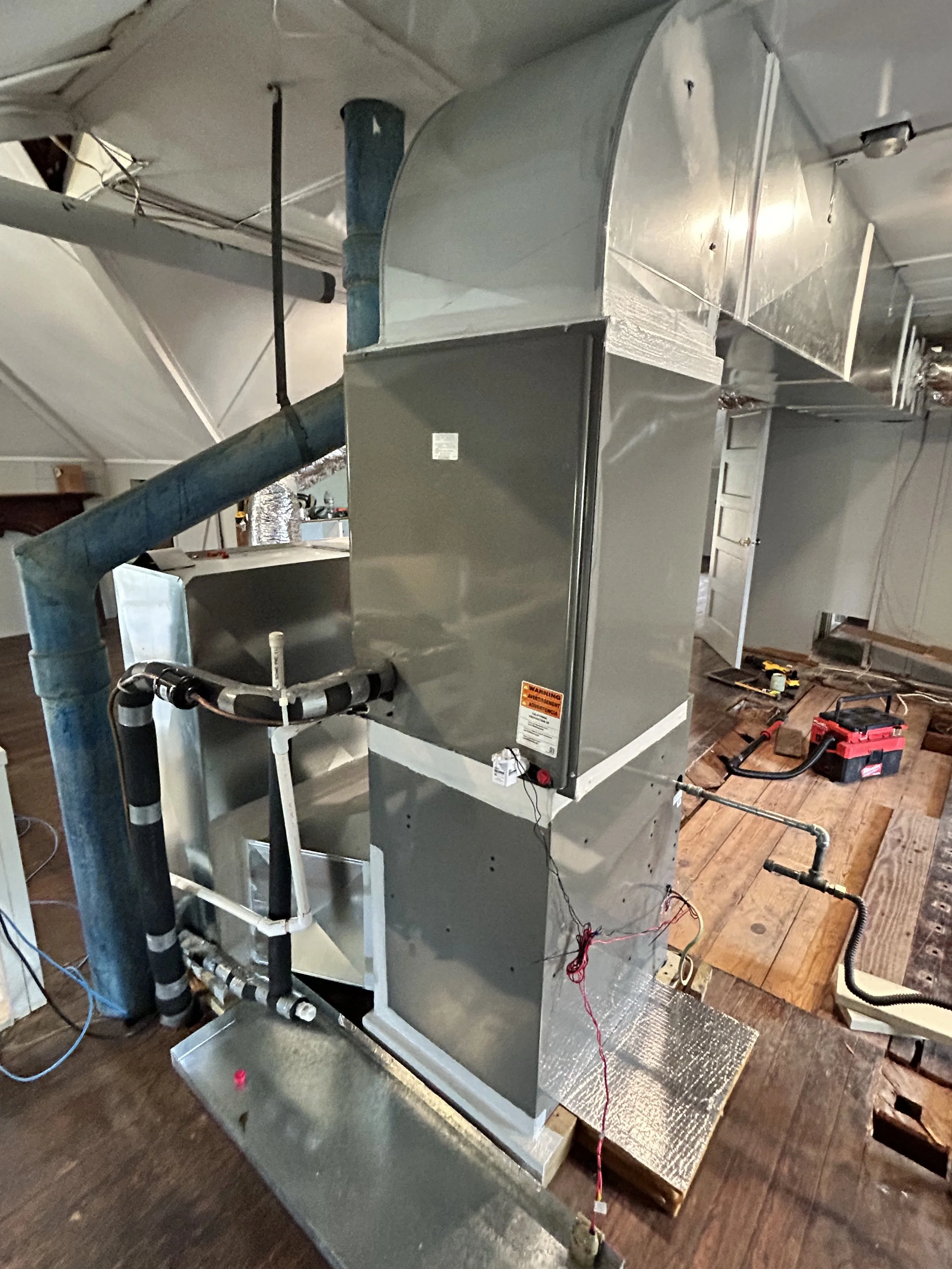 Indoor view of an HVAC system setup with metal ducts and piping, with tools and wires on the wooden floor.