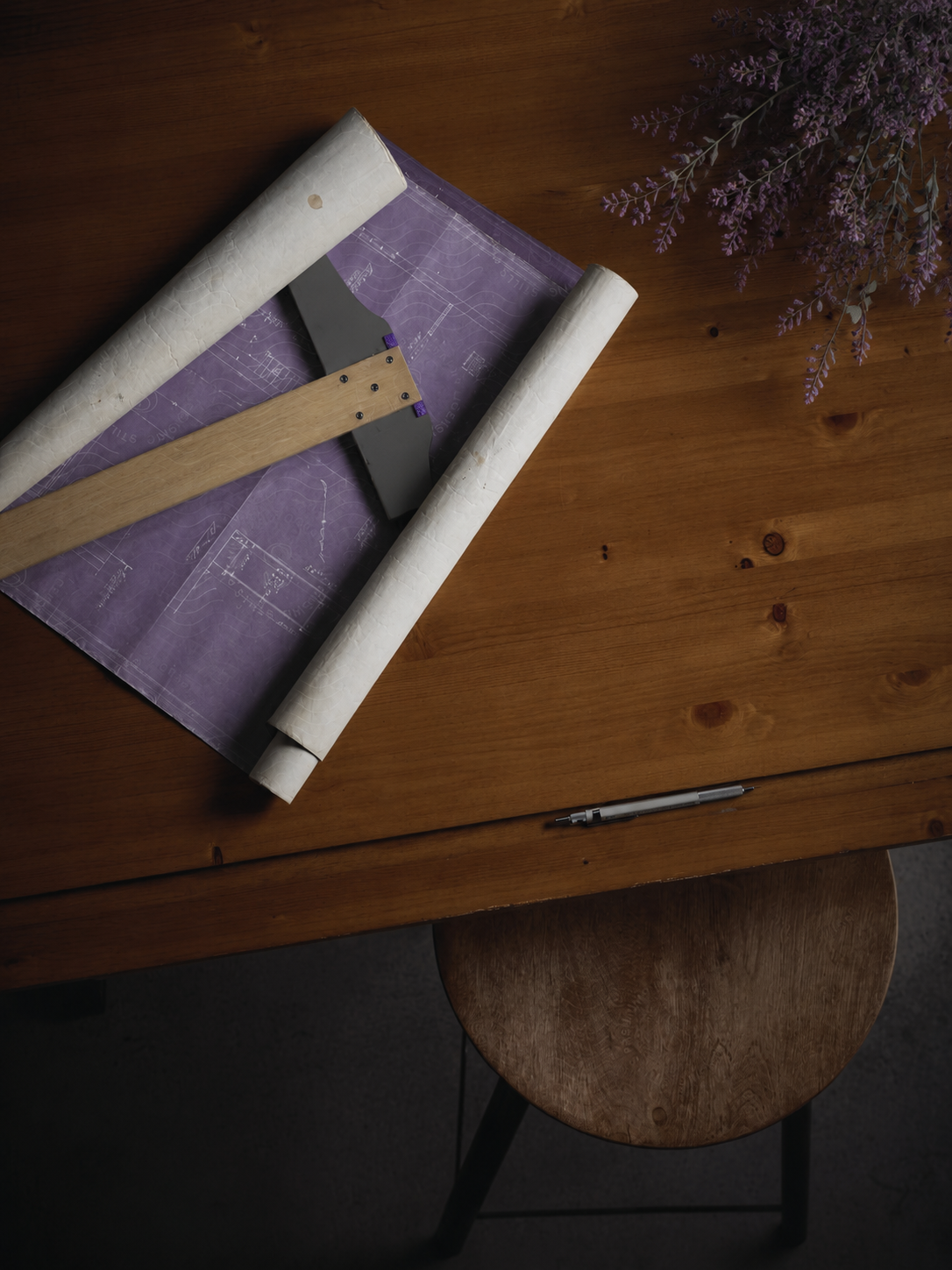 A wooden table with architectural blueprints, a folding ruler, a pen, and purple flowers in the top right corner.