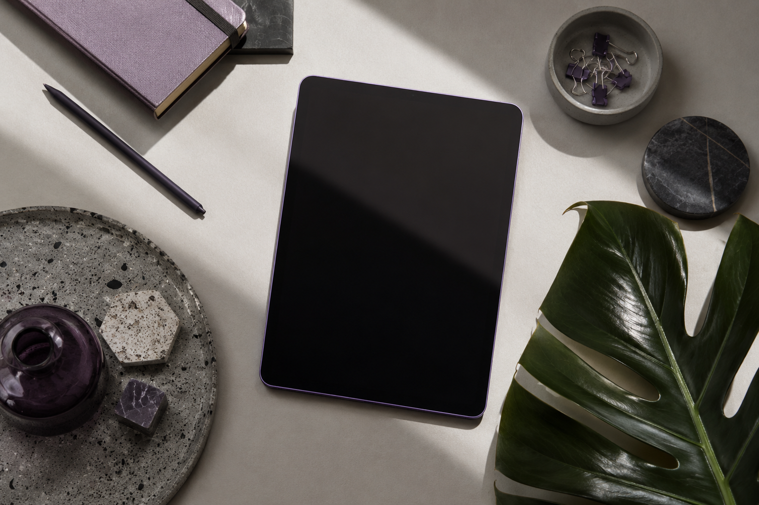 Flat lay of a workspace with a tablet, notebooks, pen, decorative items, and a large green leaf.