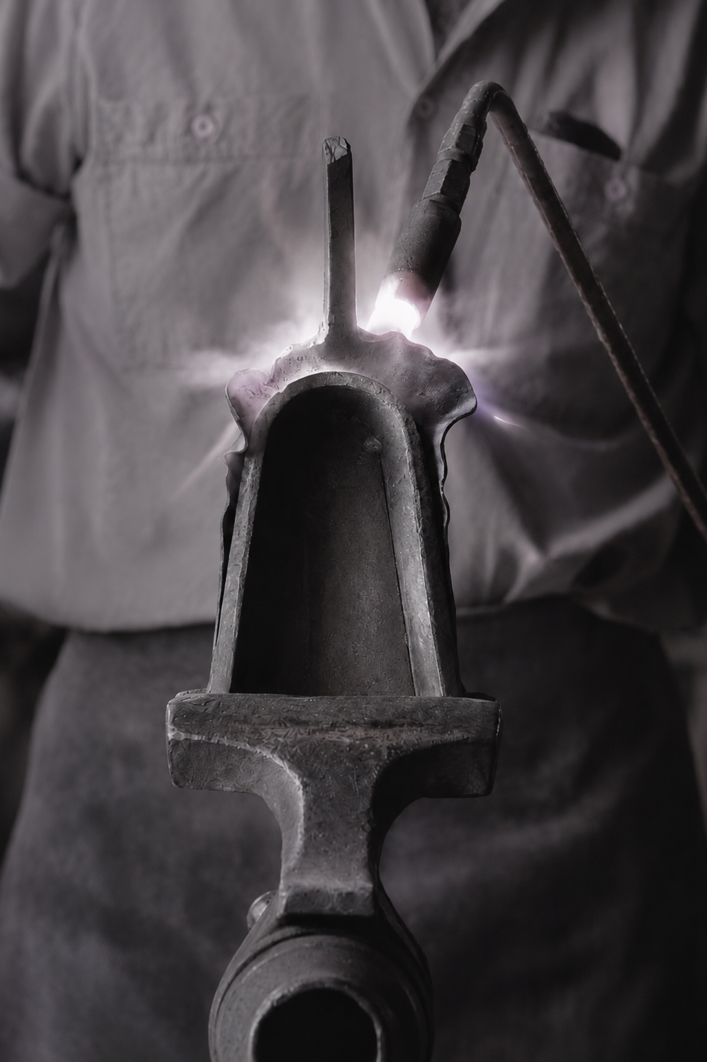 A person welding a metal pipe, with bright sparks and light emitted from the welding process, wearing a gray work shirt.