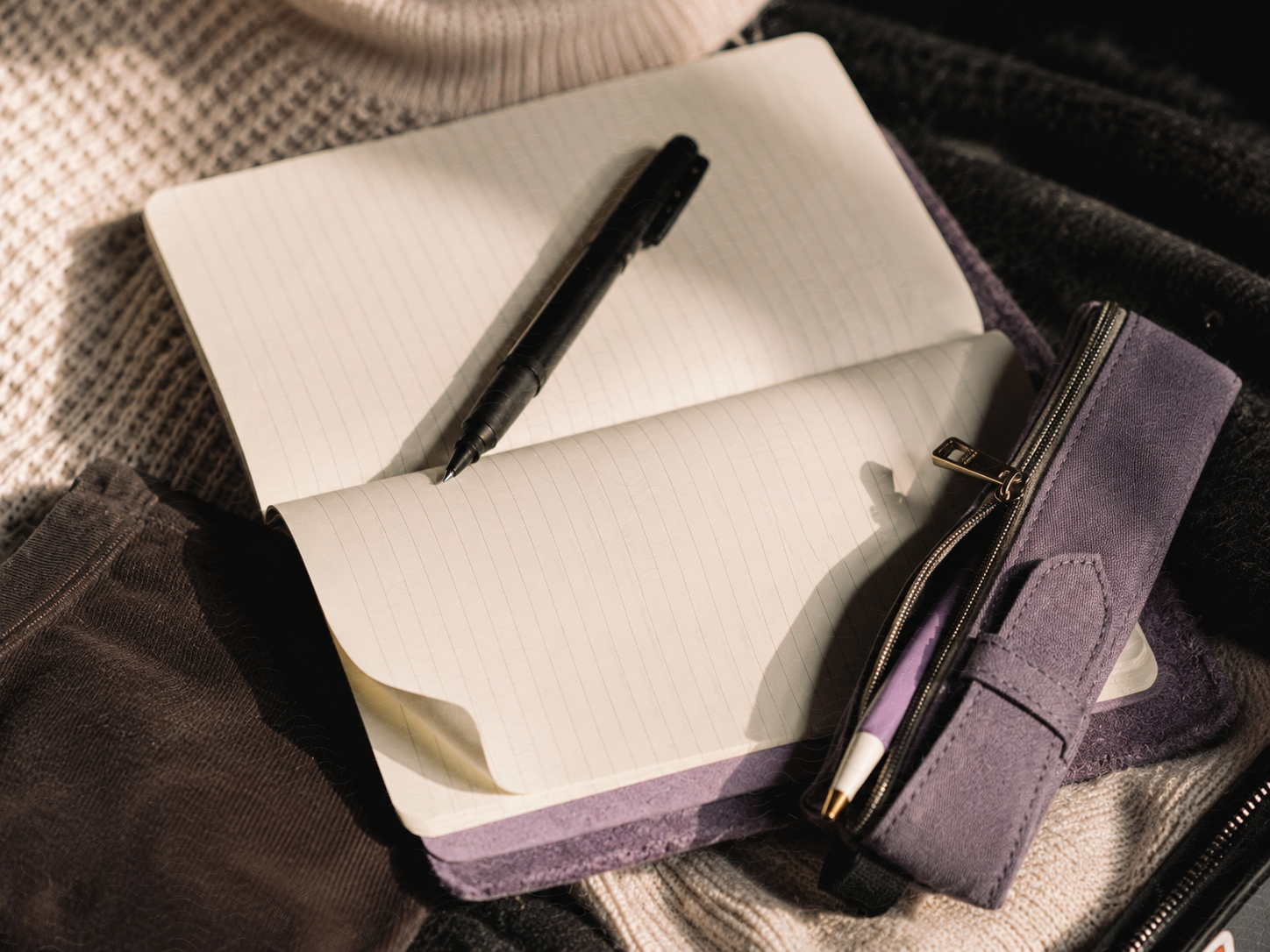 Open purple pencil case with lined notebook and pens inside, resting on textured fabric background.