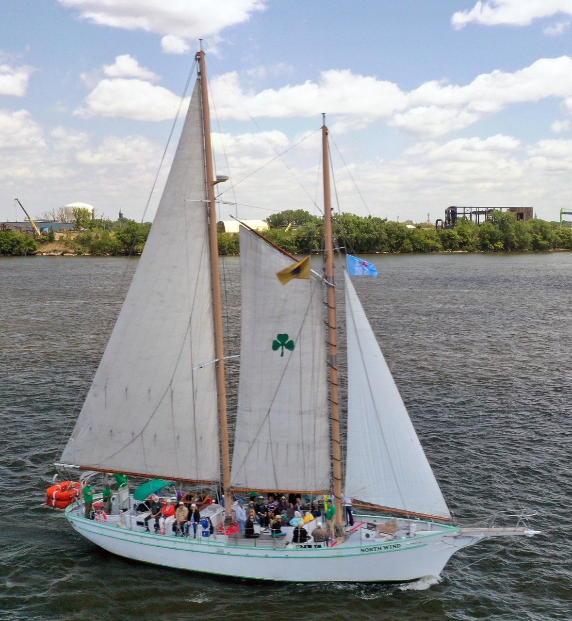 Sailboat with white sails, a green four-leaf clover symbol, and people on deck, sailing on a river with green trees and industrial buildings in the background under a partly cloudy sky.