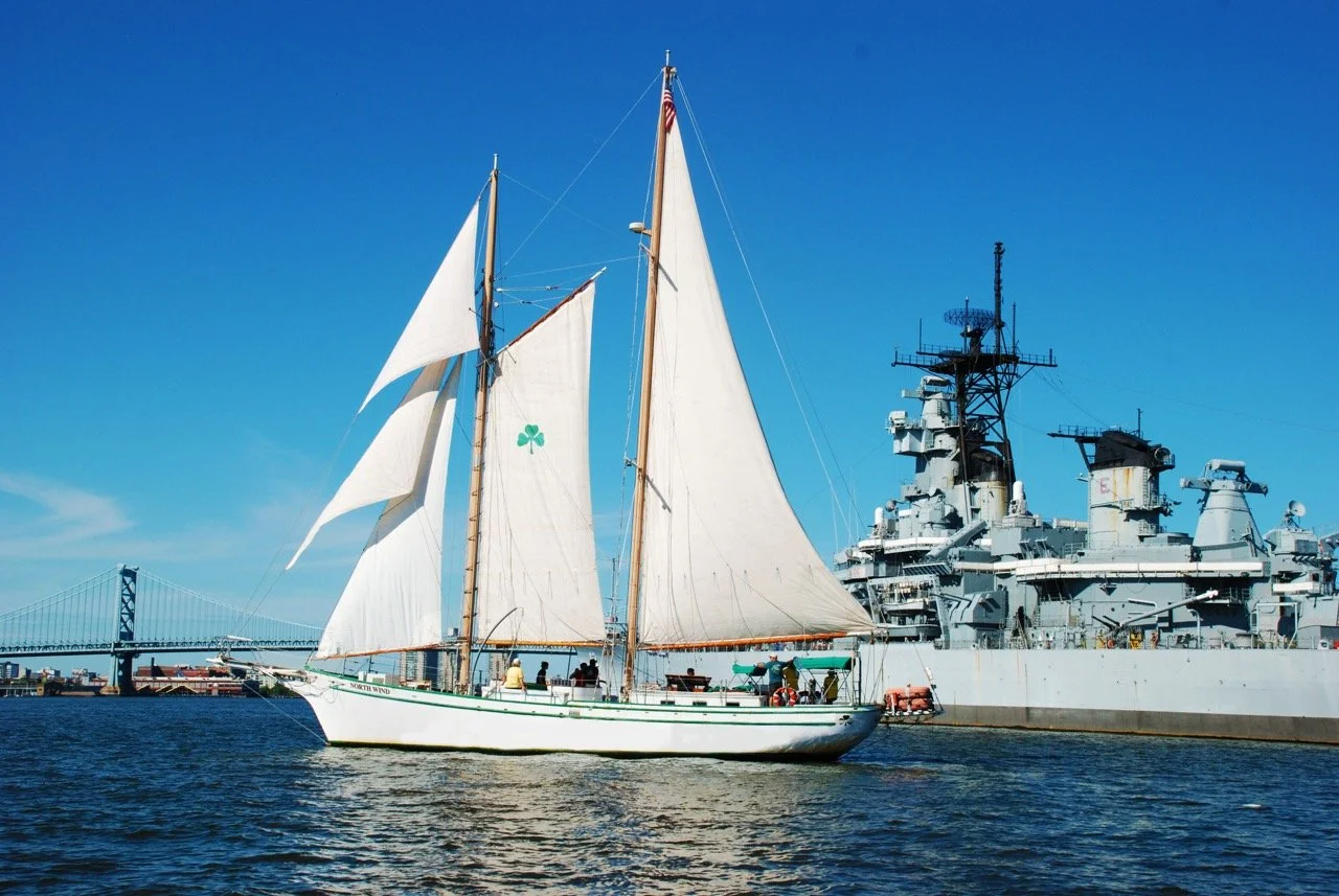 A sailboat with white sails and a green shamrock symbol on the sail, sailing near a large gray battleship, with a bridge in the background and clear blue sky.