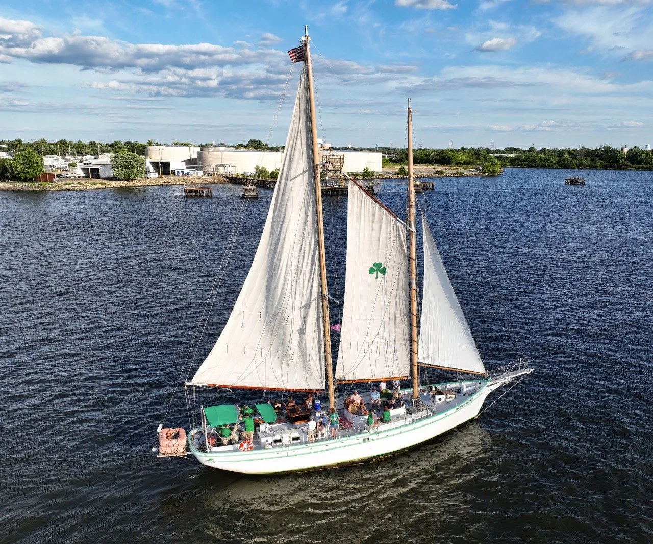 A white sailboat with two masts sailing on a body of water with people on the deck, some under a green canopy, and American flag at the top of the larger mast. The sailboat has a green shamrock emblem on one sail, and the background features a shoreline with industrial buildings and a partly cloudy sky.
