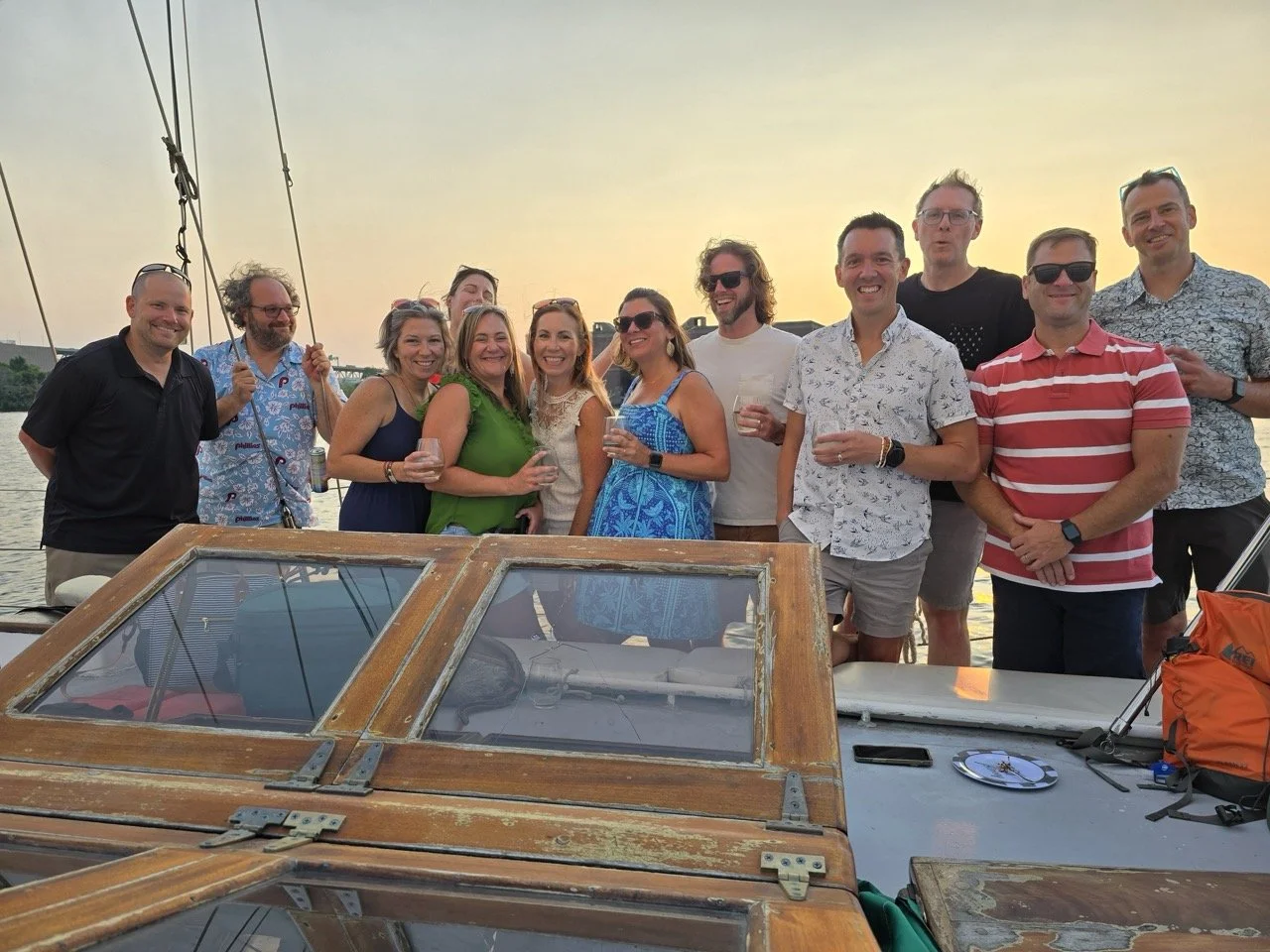 Group of people smiling and holding drinks on a boat at sunset.
