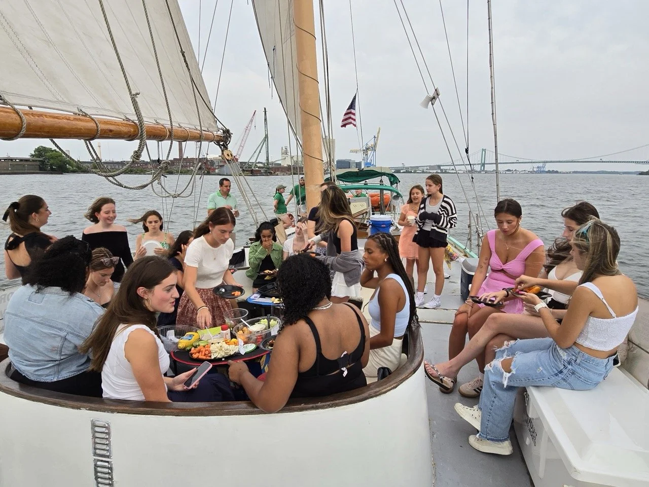 A group of people enjoying a gathering on a sailboat on the water, with some preparing food and others socializing, under an overcast sky.