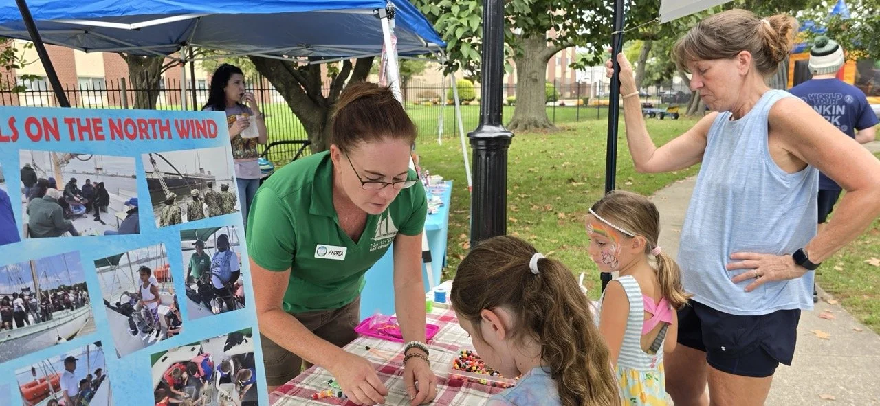 Two women, one wearing a green shirt and glasses and the other in a blue sleeveless top, are at an outdoor booth with children. The children have face paint, and one girl is looking at beads on a table. The booth has a display with photos of people on boats, and there is a park with trees and a sidewalk in the background.