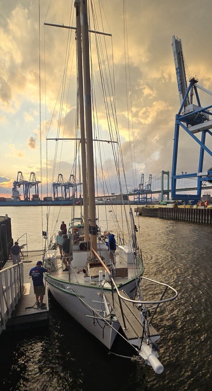 A sailboat docked at a marina during sunset with people on board and on the dock, cranes and industrial structures in the background, and a cloudy sky.