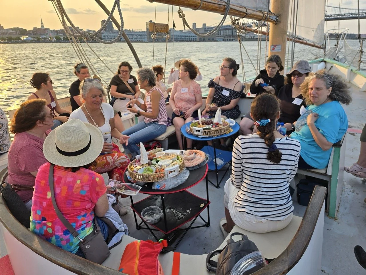 Group of women enjoying a meal together on a boat during sunset, with water and city skyline in the background.