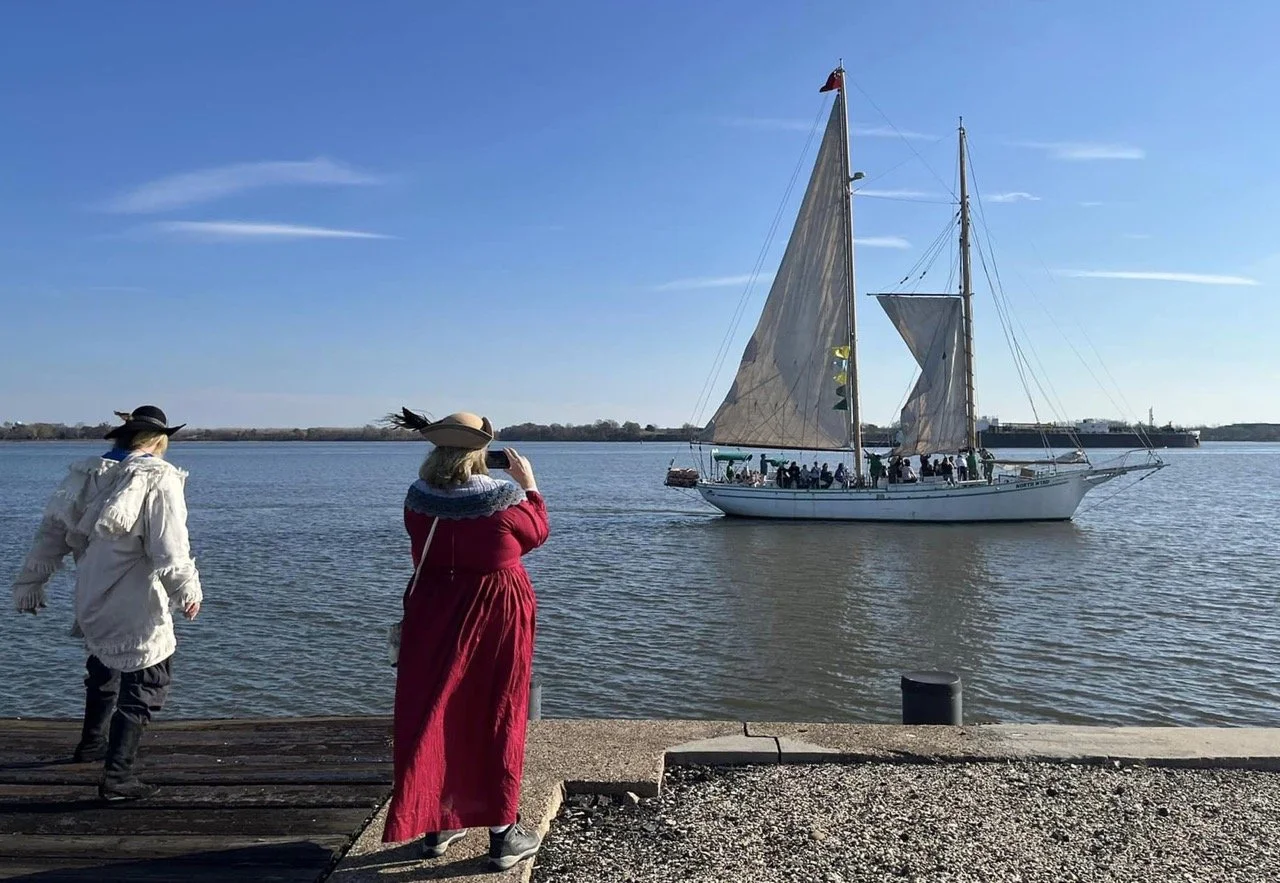 People on a dock watching a large sailboat on the water with blue sky and a few clouds.