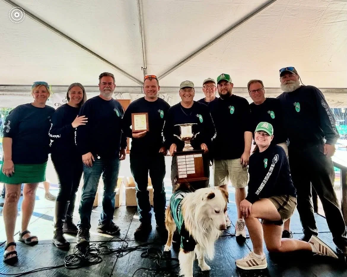 A group of nine people standing on a stage inside a tent, holding awards and plaques. A dog wearing a harness is in front of them. The group is smiling and appears to be celebrating an achievement.
