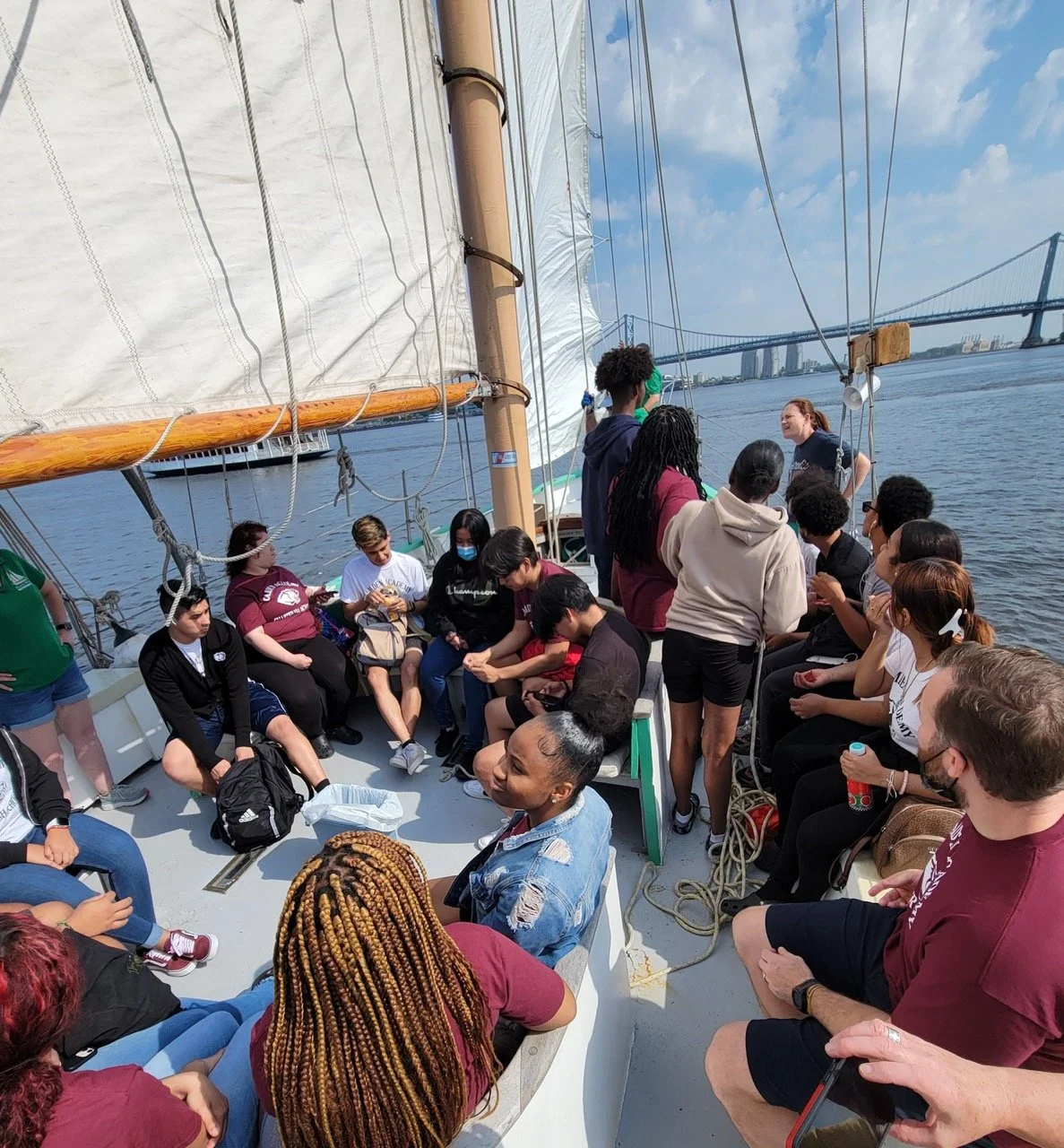 A group of people on a sailboat, some seated and others standing, with a bridge and water in the background.