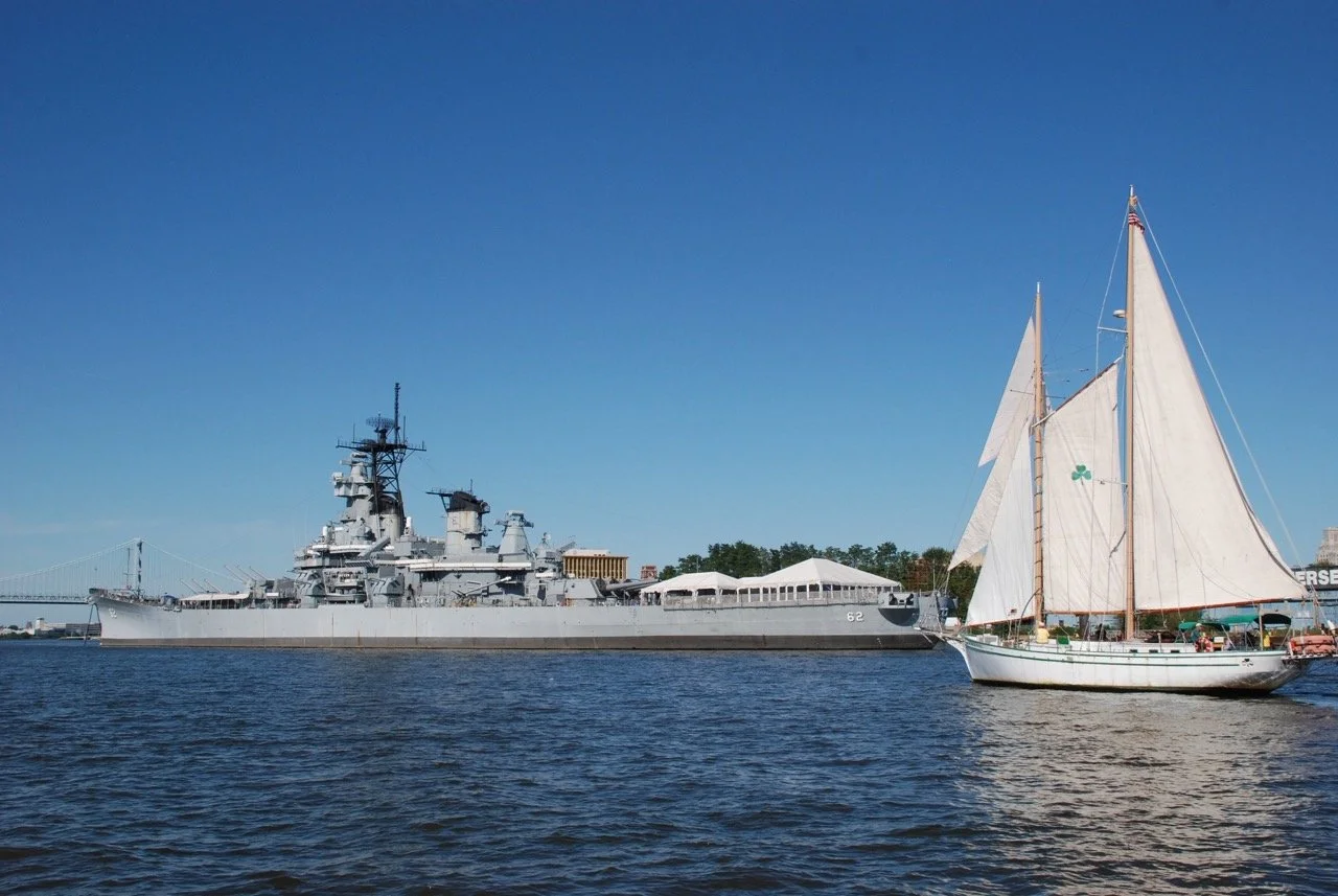 A large gray battleship sailing on water with a smaller white sailboat nearby, under a clear blue sky.