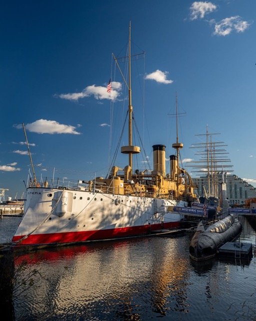 Historic warship docked at a marina during daytime with a partly cloudy sky.