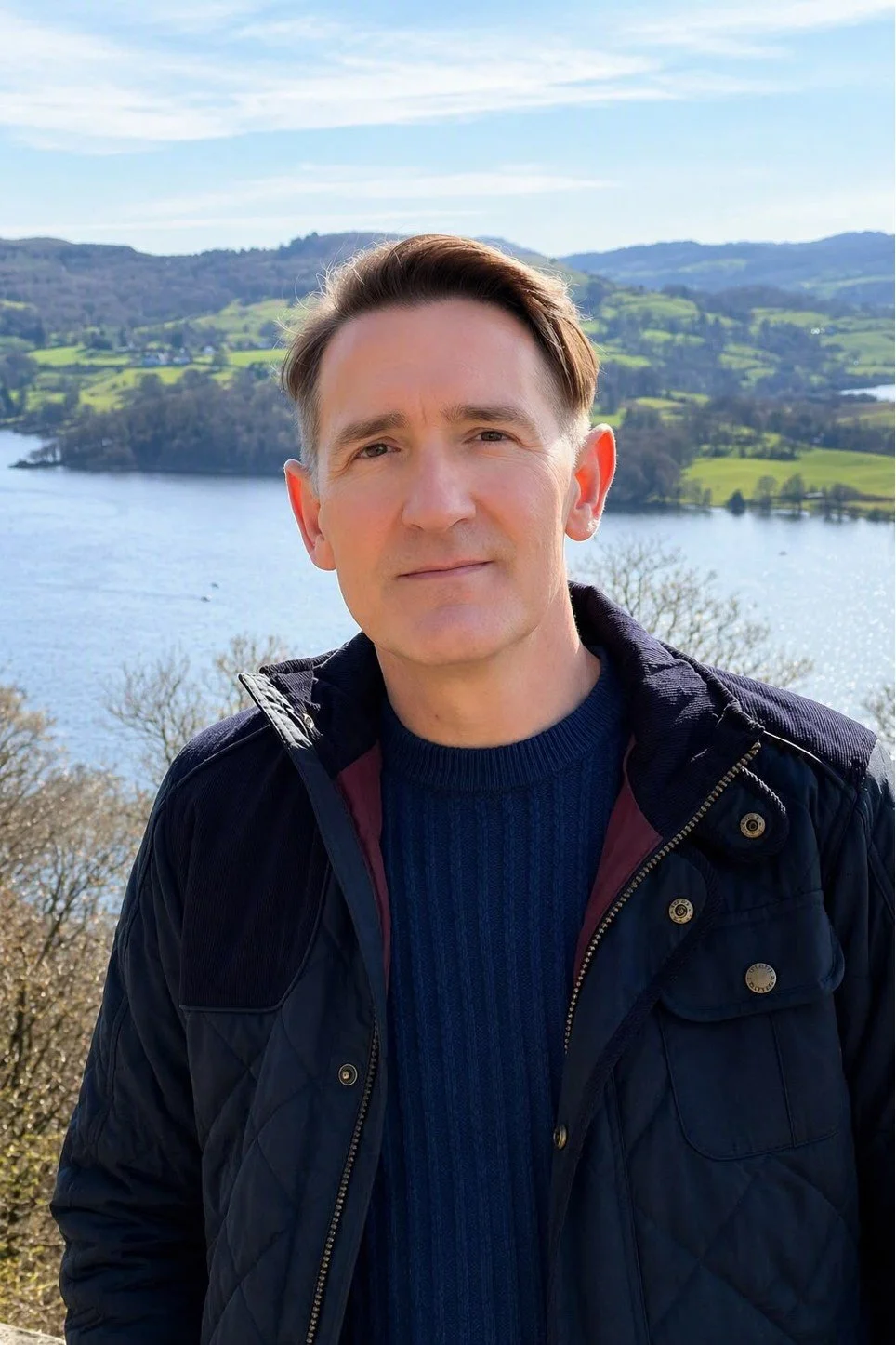 A man standing outdoors by a lake, with rolling green hills and trees in the background, wearing a dark jacket and sweater.