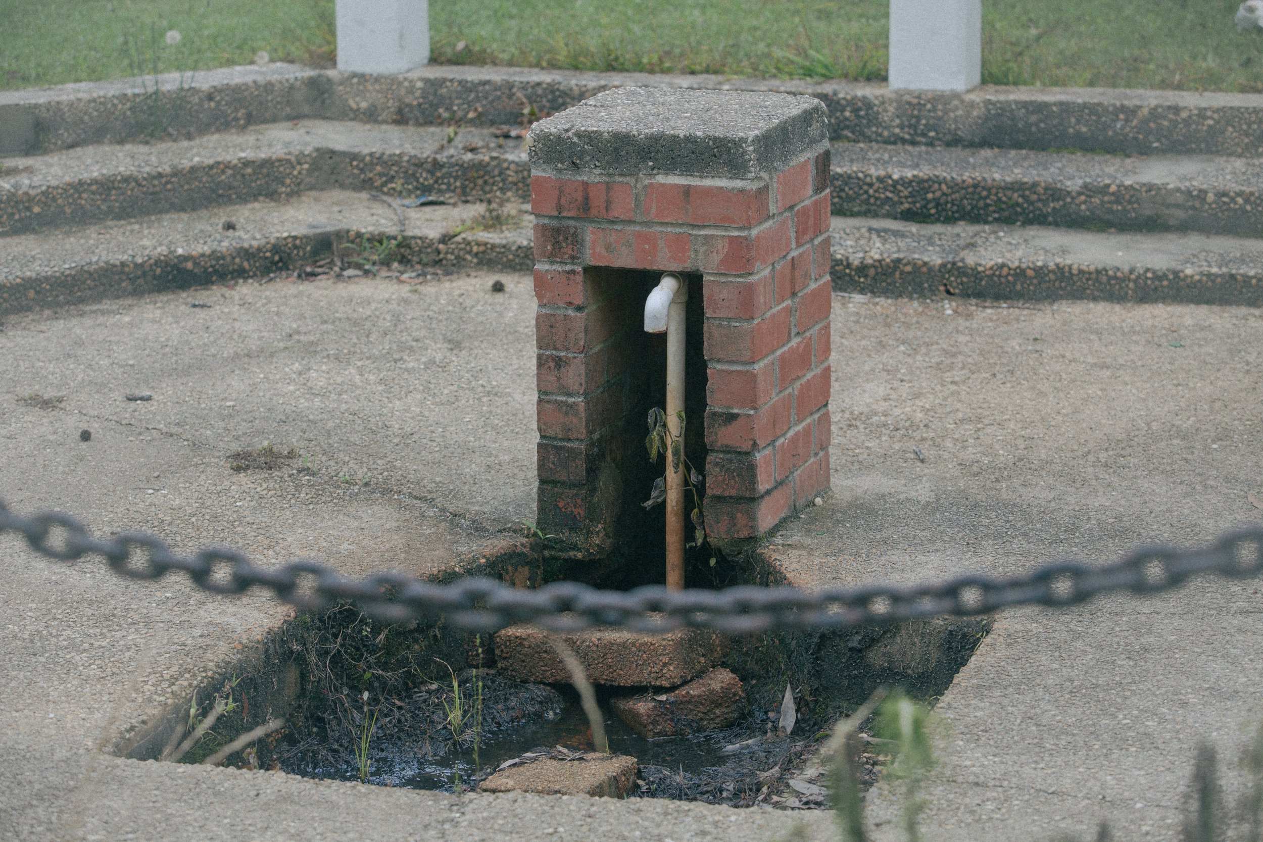 A small brick structure covering a pipe, with a hole underground and a flat top, situated on a concrete sidewalk with grassy area in the background.