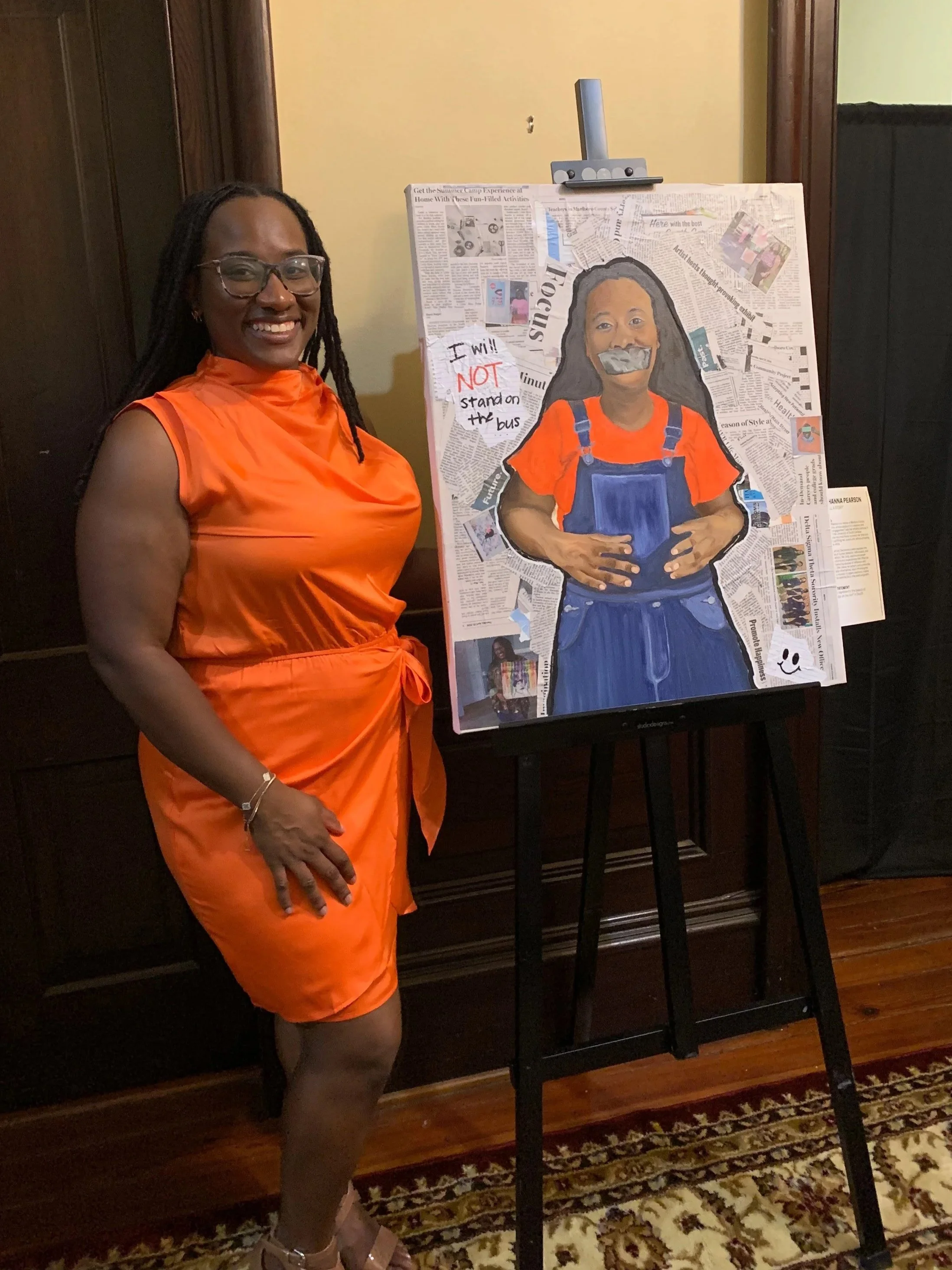 A woman with dark skin, wearing glasses and a bright orange dress, standing next to a collage art piece with newspaper clippings and a painted portrait of a woman with long black hair, wearing a red top and blue overalls with tape over her mouth, on an easel in a room with wood floors and decorated rug.