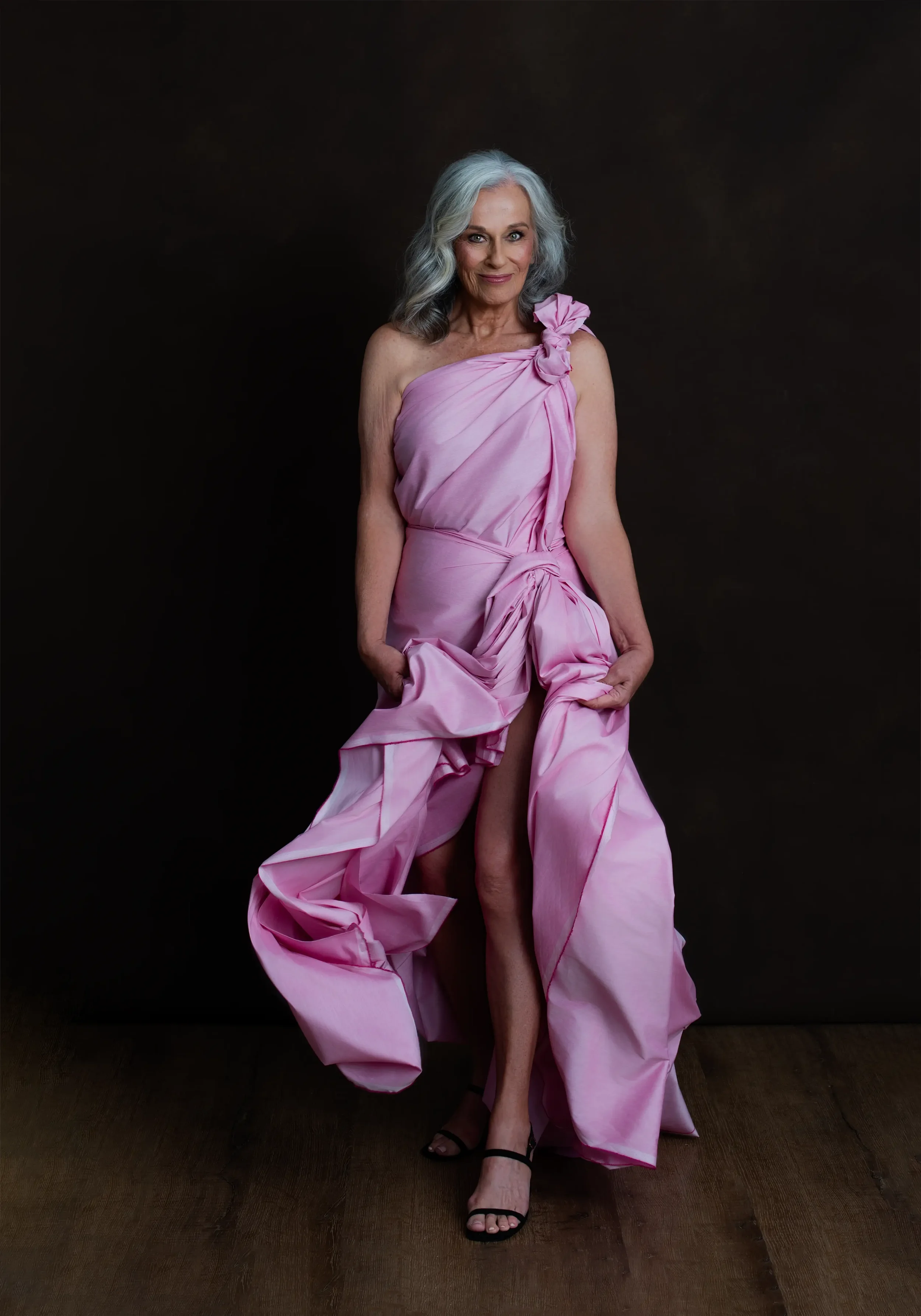An older woman with gray hair wears a pink gown with ruffles and bows, standing against a dark background. She is smiling and looking at the camera, holding the gown to reveal her legs.
