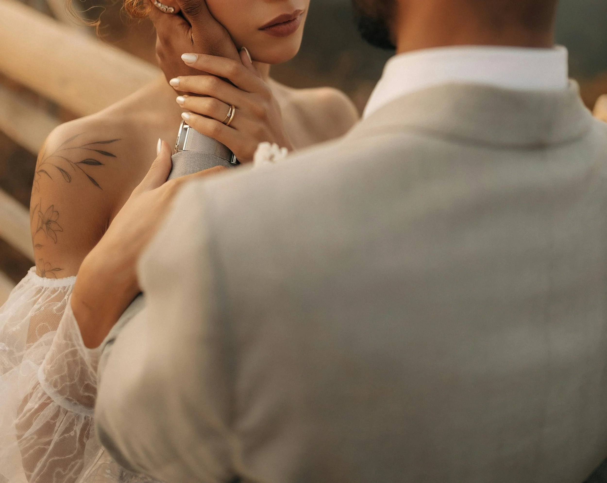 Close-up of a woman with a tattoo on her shoulder, touching her face, wearing pearl earrings, and holding a wristwatch. She is dressed in lace and is near a man in a white shirt.