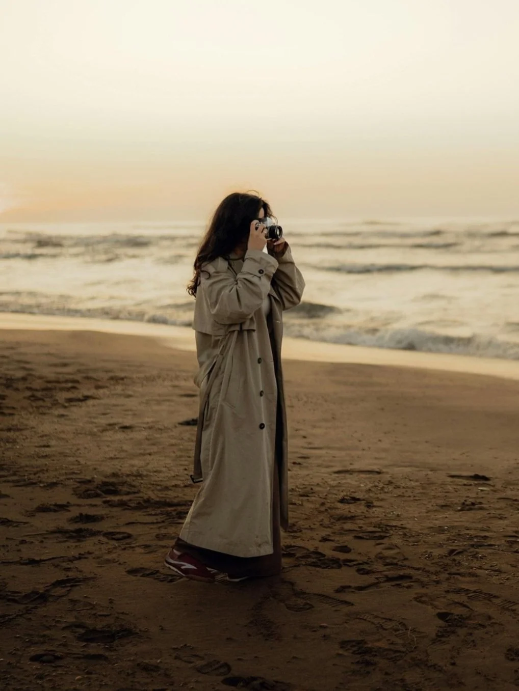 A woman with long dark hair wearing a tan trench coat and brown shoes standing on a sandy beach, taking a photograph with a camera during sunset.