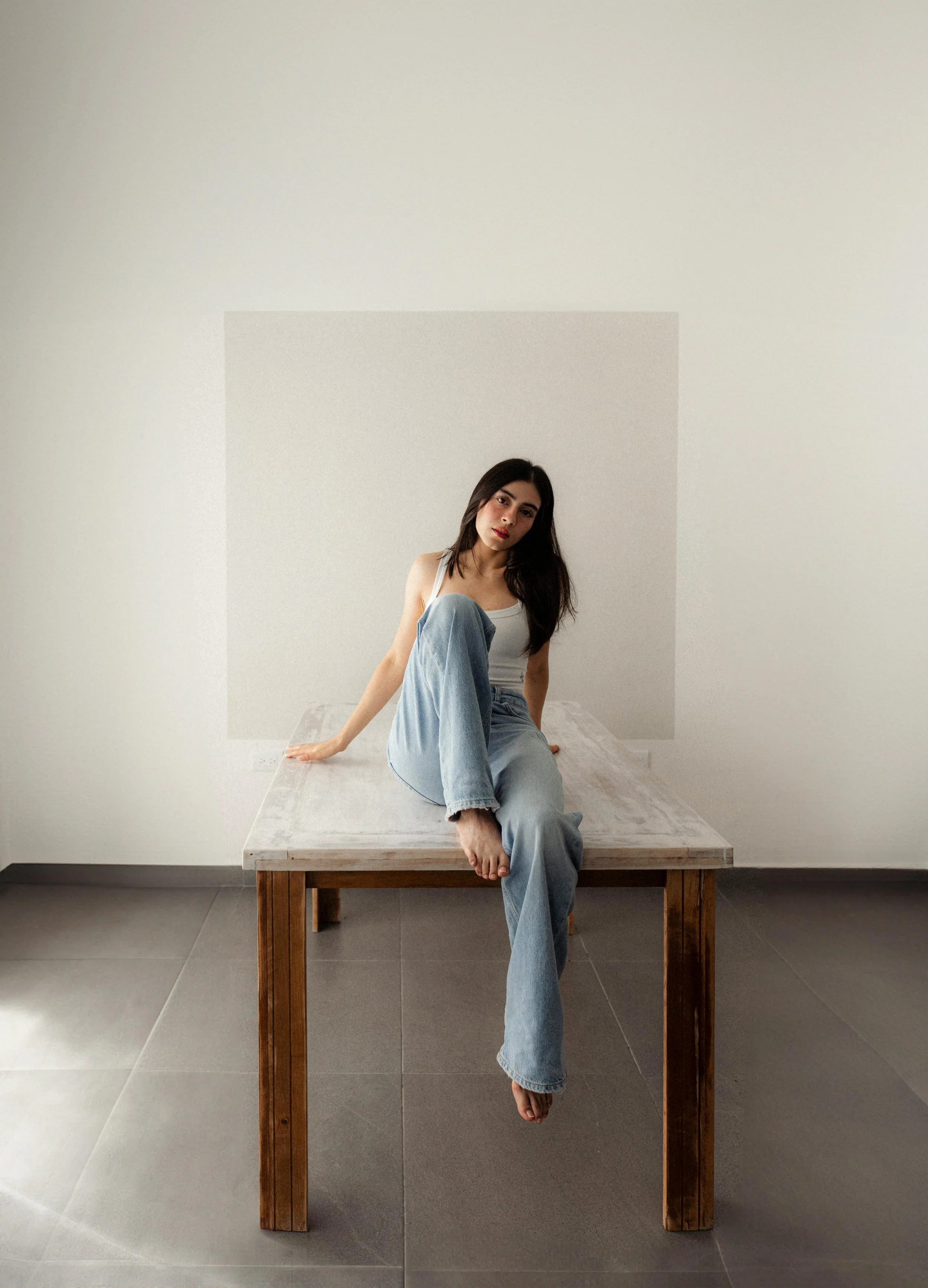 A young woman with long dark hair sitting on a wooden table in front of a plain white wall with a small square painted section behind her. She is wearing a white tank top and loose blue jeans, with one leg bent and foot resting on the table, looking into the camera.
