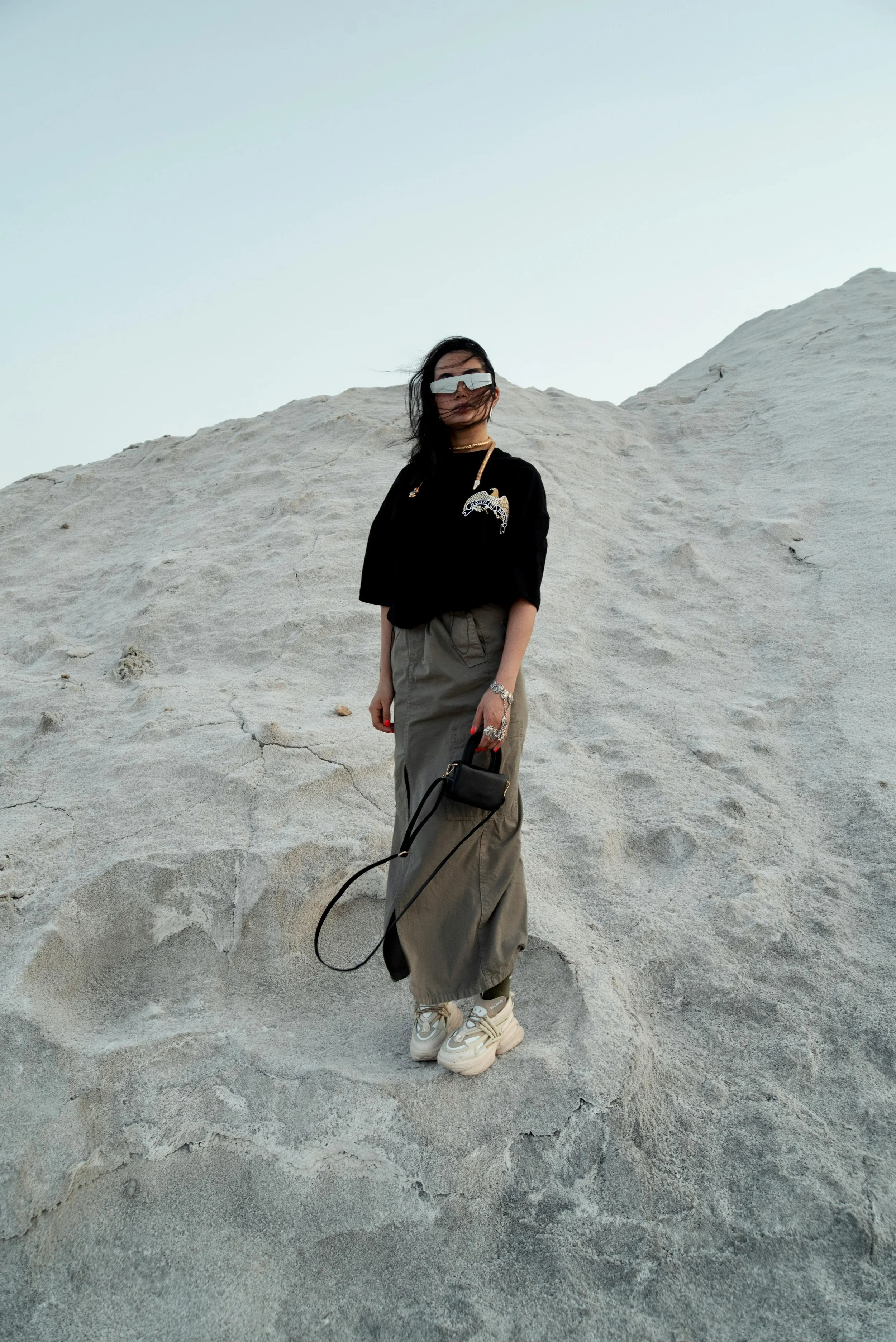 A woman standing on a sandy, cracked surface that appears to be a desert or salt flat, wearing a black shirt, long gray skirt, chunky sneakers, mirrored sunglasses, and holding a small black handbag.