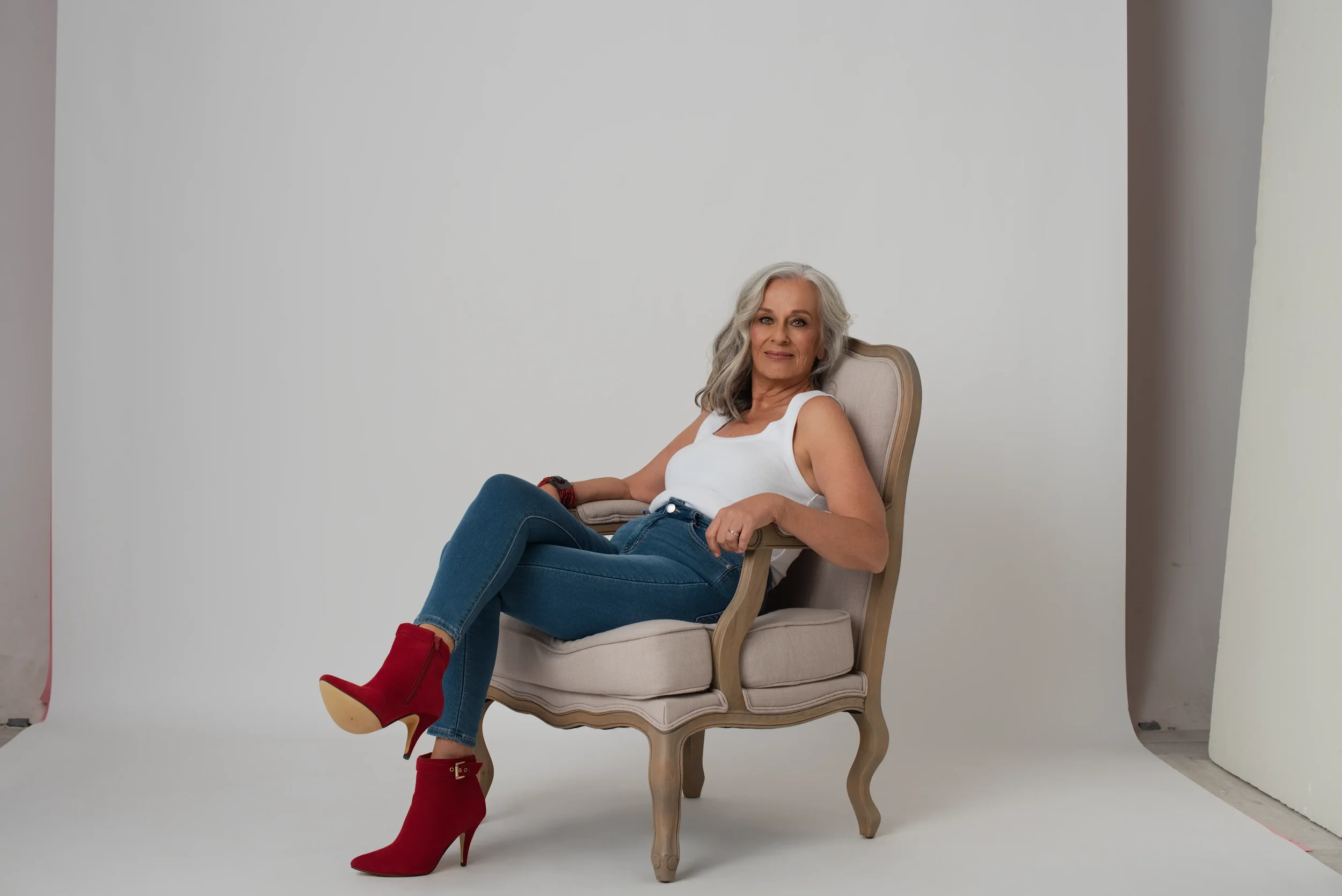 Silver hair lady wearing jeans, white tank top and red ankle boots. She is sitting on a chair on a white paper background in a women's portrait photo studio.