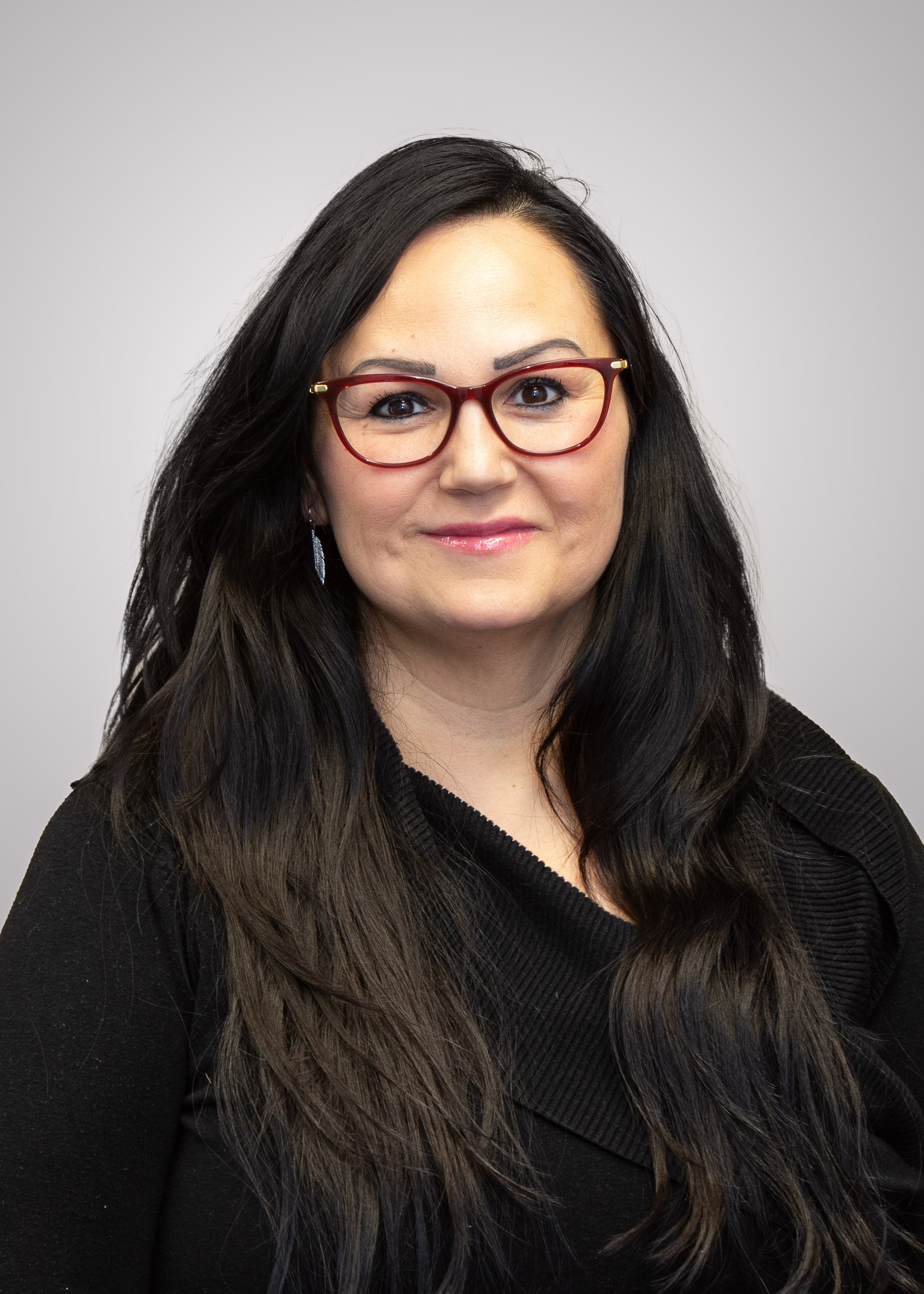 A woman with long dark hair, wearing red glasses and a black top, smiling at the camera against a plain light gray background.