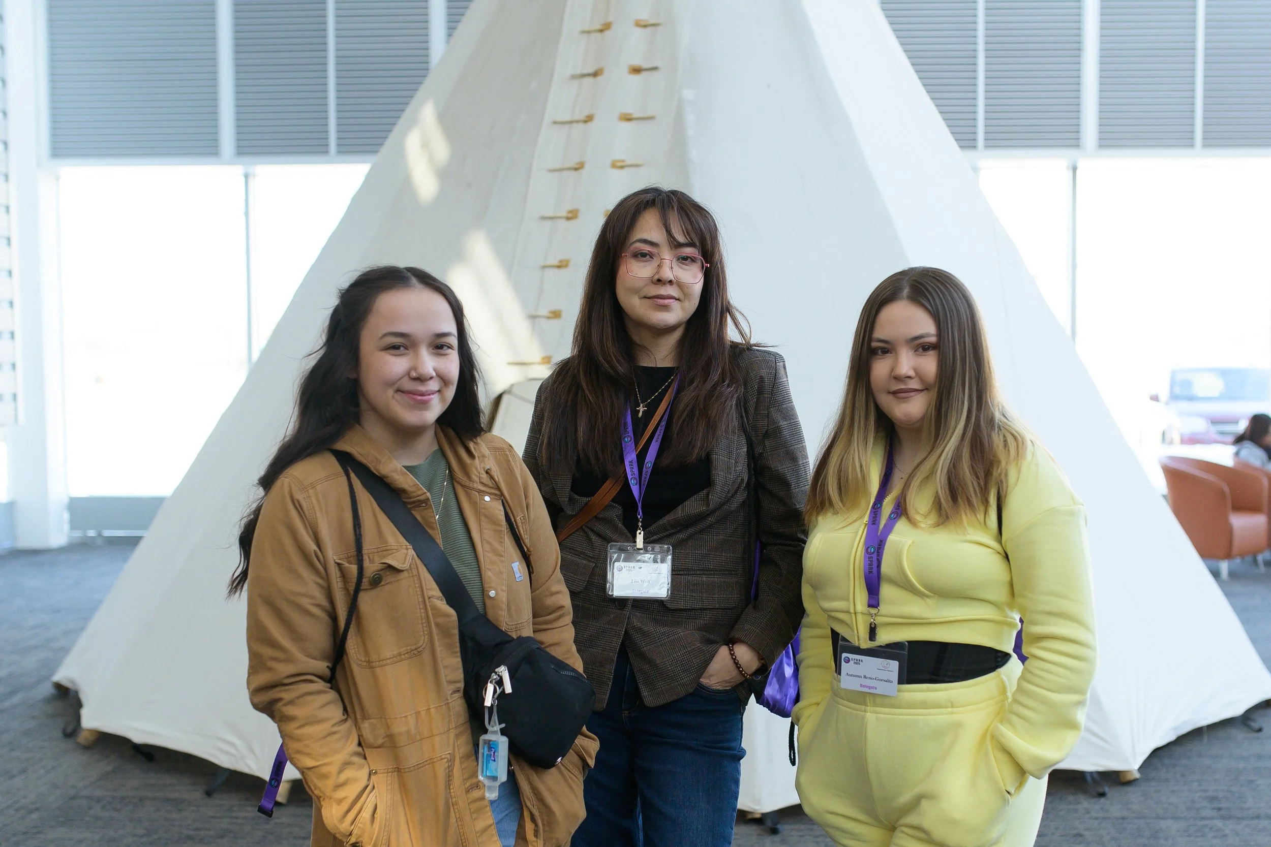 Three women standing together at an indoor event, wearing name badges and lanyards, with a white tent-like structure in the background.