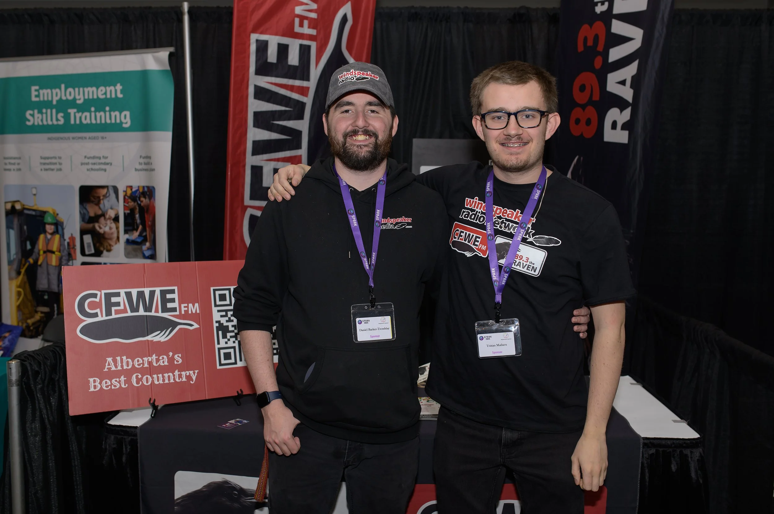 Two young men standing side by side at an event, smiling at the camera. They are both wearing black shirts with various patches and lanyards with badges. The background has black curtains, banners, and a promotional display for a radio station.
