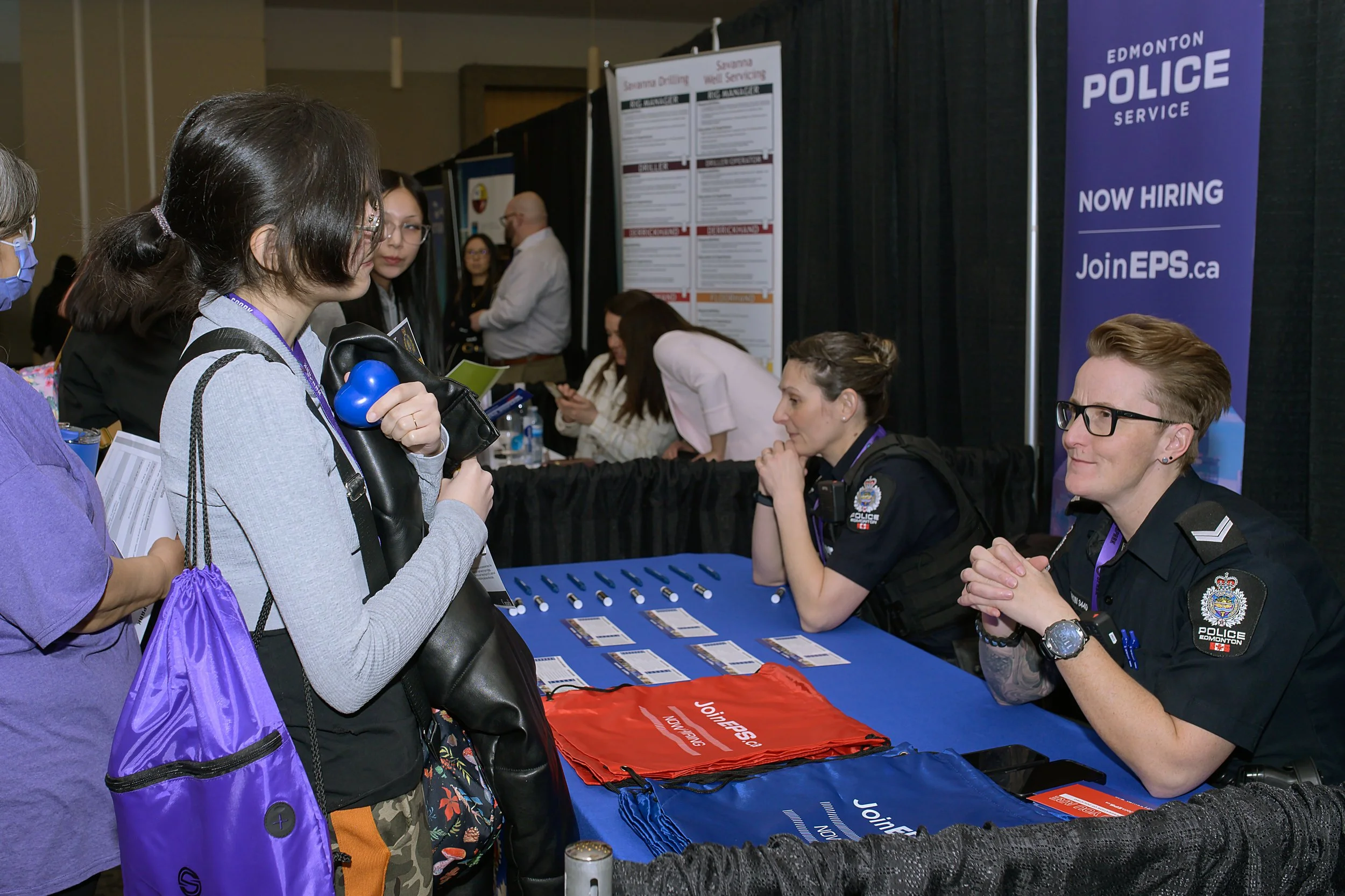 A career fair booth for Edmonton Police Service, with officers sitting behind a table displaying safety pins and promotional materials, interacting with attendees.