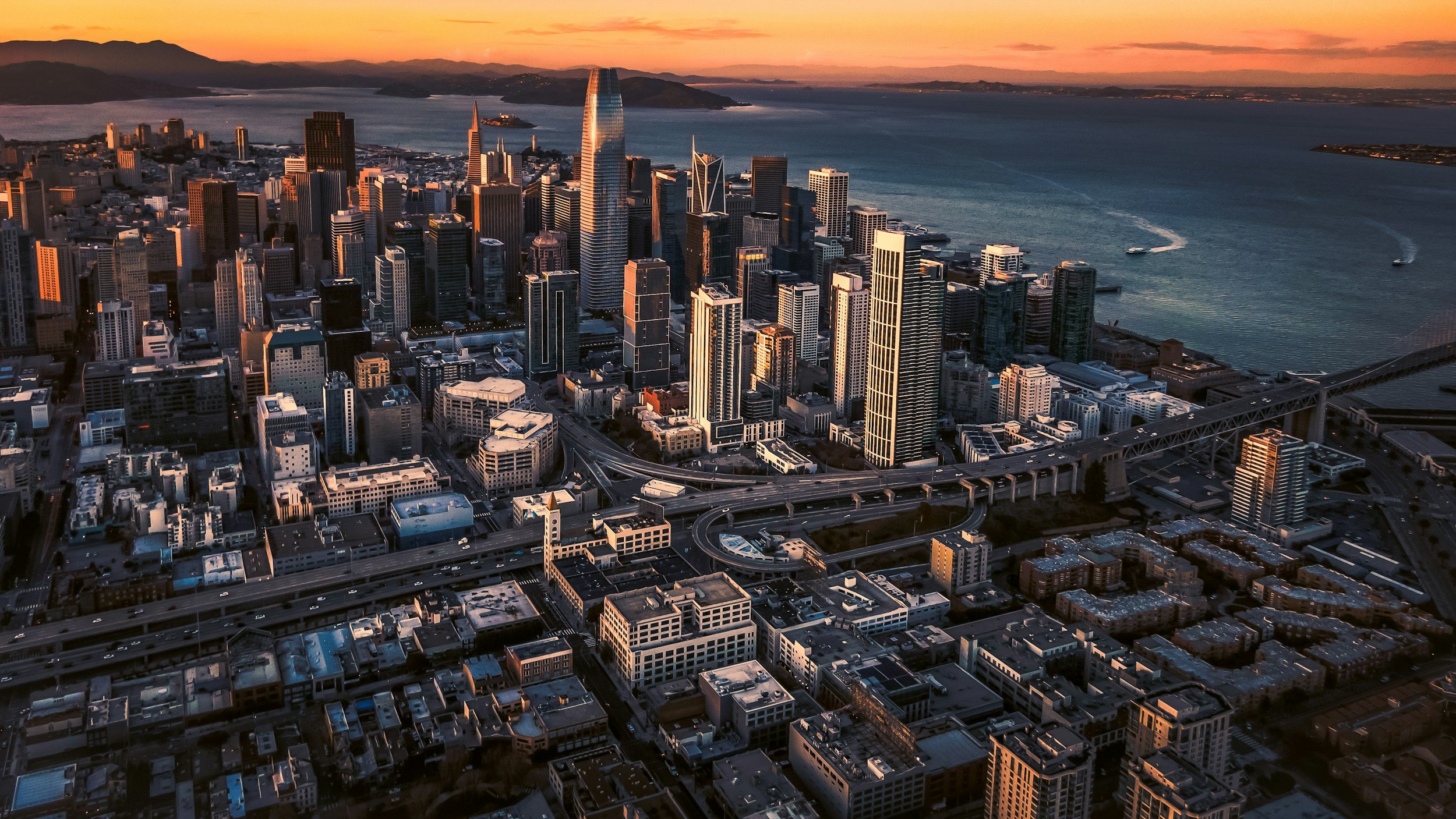 Aerial view of downtown San Francisco at sunset, showing tall skyscrapers, bridges, and the bay with boats and hills in the background.