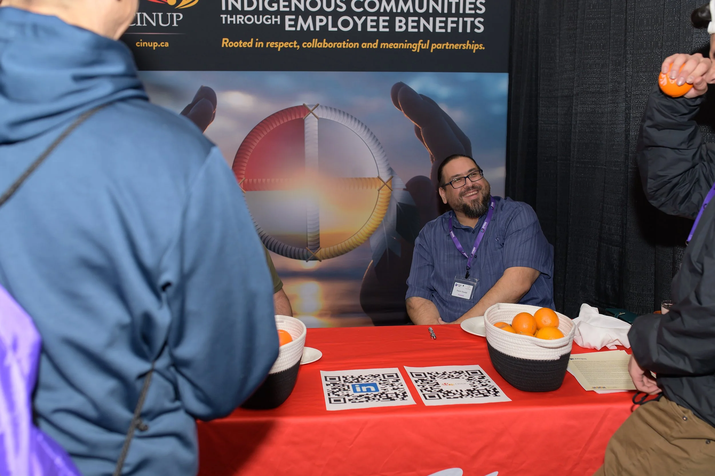 People gathered around a booth table with baskets of oranges, QR codes, and informational materials, in front of a large poster about Indigenous communities and employee benefits.