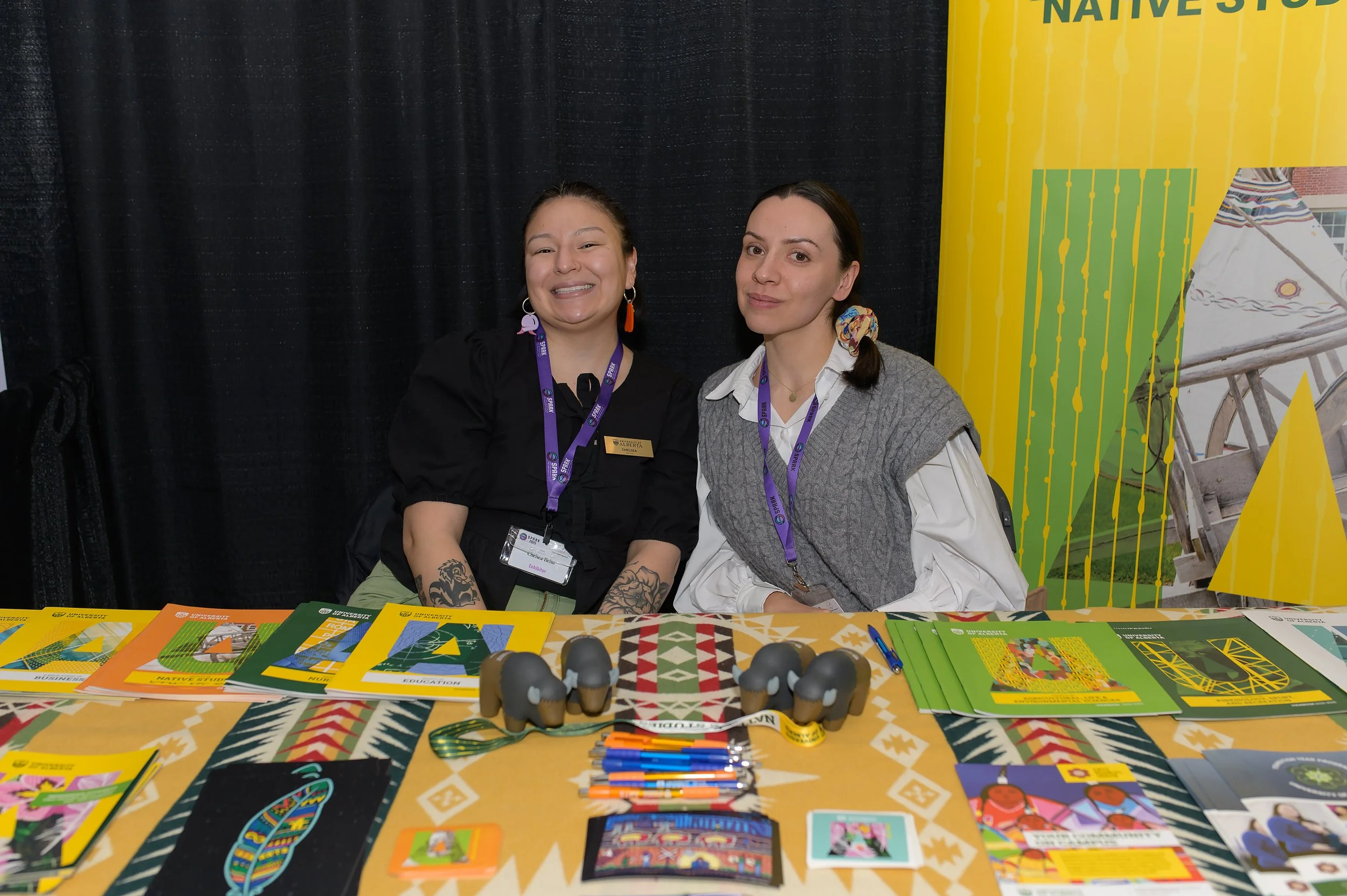 Two women sitting at a booth with colorful brochures, pens, and small figurines, with a black backdrop and yellow booth display behind them.