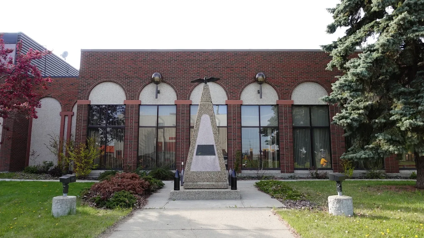 Memorial monument in front of a brick building, with a triangular obelisk, plaques, two eagle sculptures on top, and surrounding greenery.