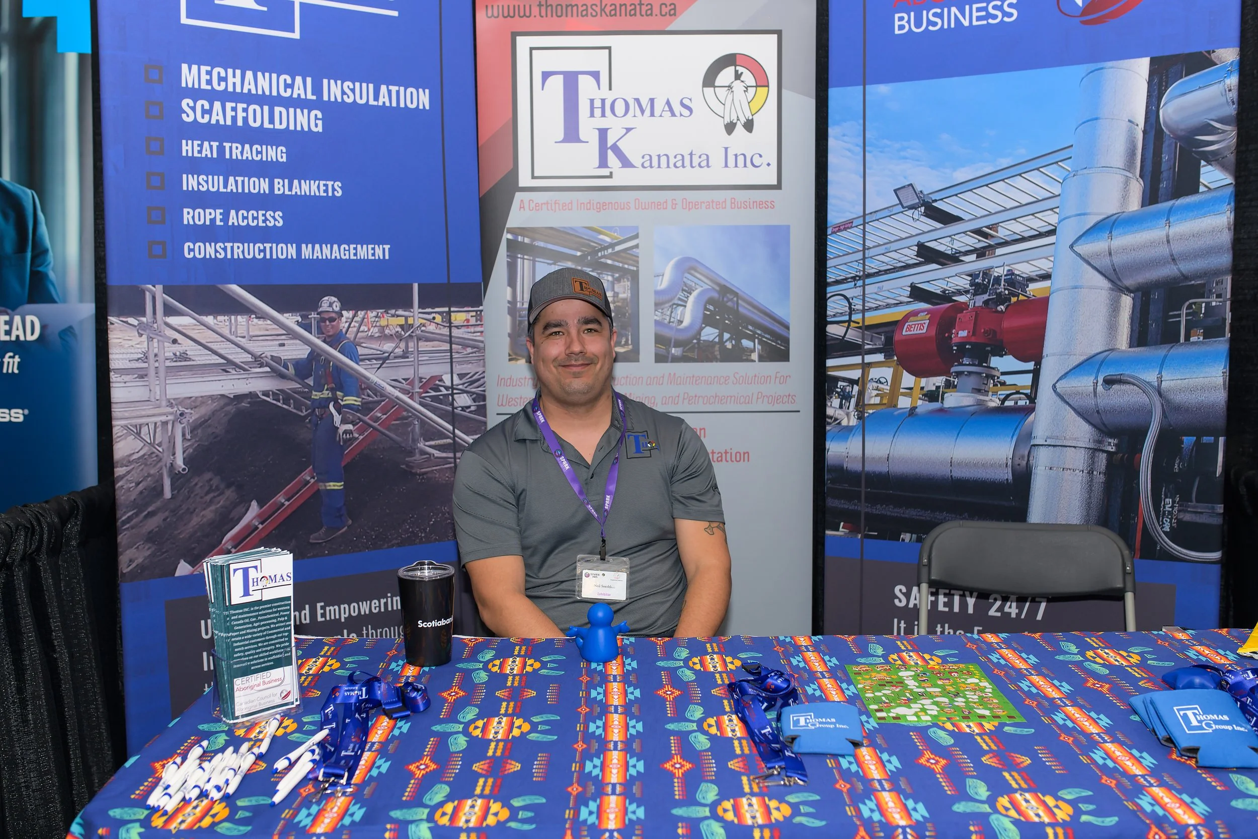 A man sitting at a booth with a colorful tablecloth, promotional materials, and blue and white pens at an outdoor event. Behind him are banners for Thomas Kanata Inc., a business specializing in mechanical insulation, scaffolding, heat tracing, insul