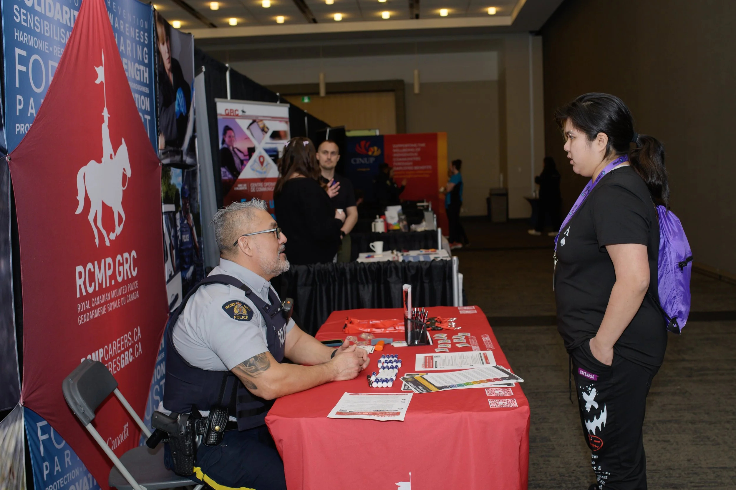 A man in a police uniform sitting at a booth, speaking with a woman standing in front of him at a conference. The booth has a large red banner with a white logo of a horse and officer with text related to Royal Canadian Mounted Police. The table has 