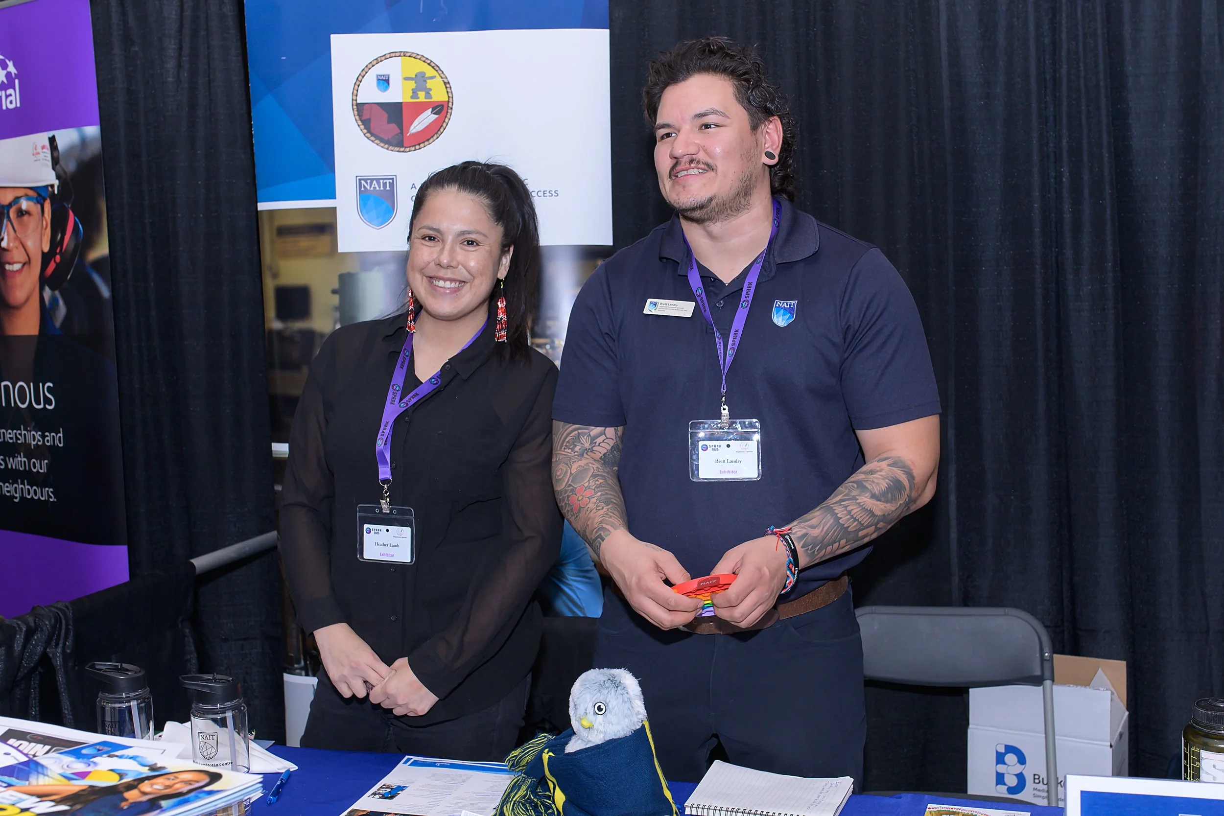 Two individuals standing behind a table at an event, both wearing conference badges and lanyards. The woman on the left has dark hair and is smiling, while the man on the right has tattoos on his arms, dark curly hair, and is also smiling. There are 