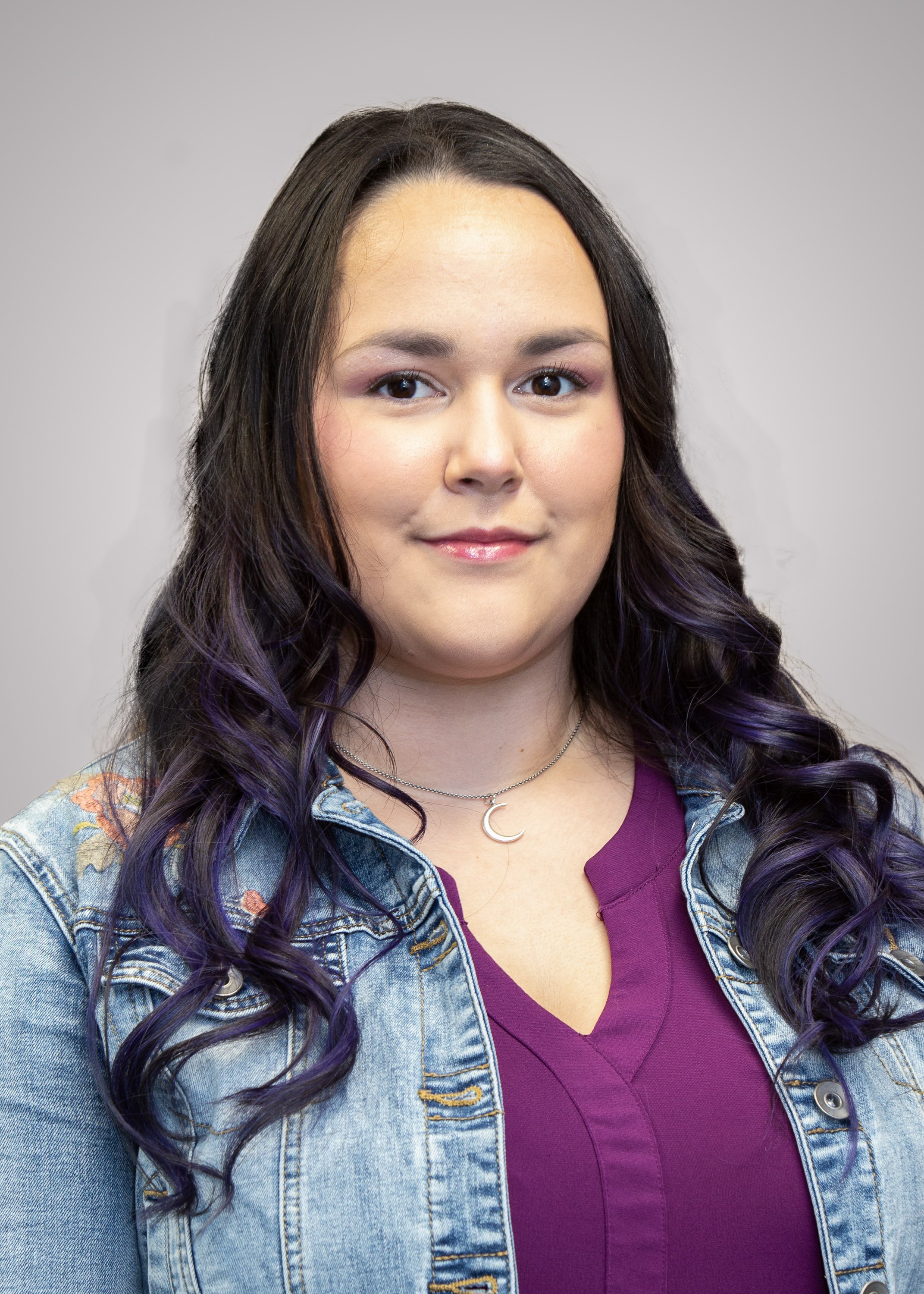 A young woman with long, wavy hair dyed purple at the ends, wearing a denim jacket, a purple top, and a moon-shaped pendant necklace, smiling against a plain gray background.
