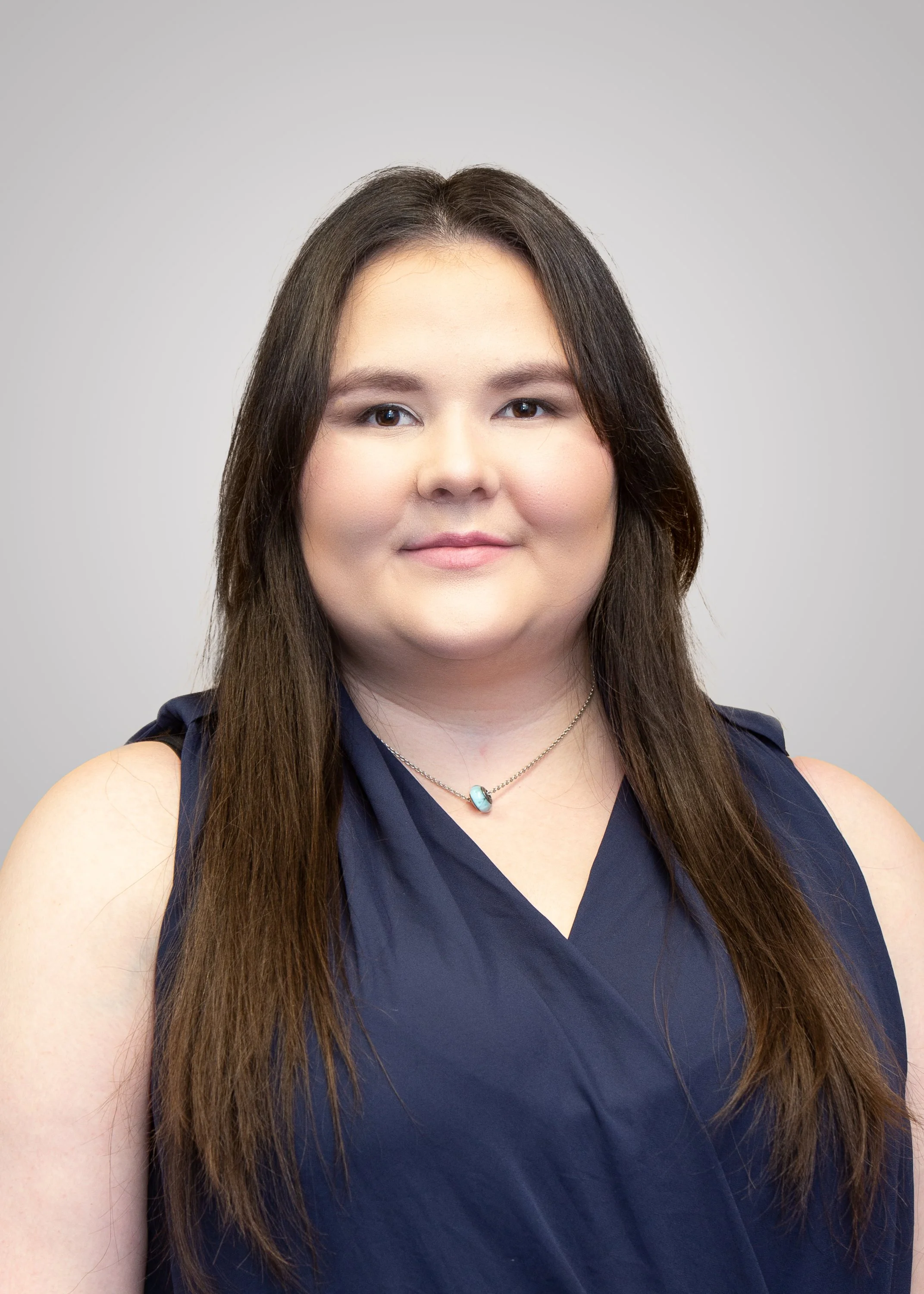 A woman with long brown hair wearing a sleeveless navy blue top and a necklace with a blue pendant, smiling softly against a plain gray background.