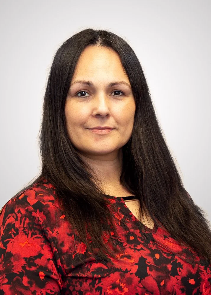 Portrait of a woman with long dark hair wearing a red and black patterned top against a plain light background.