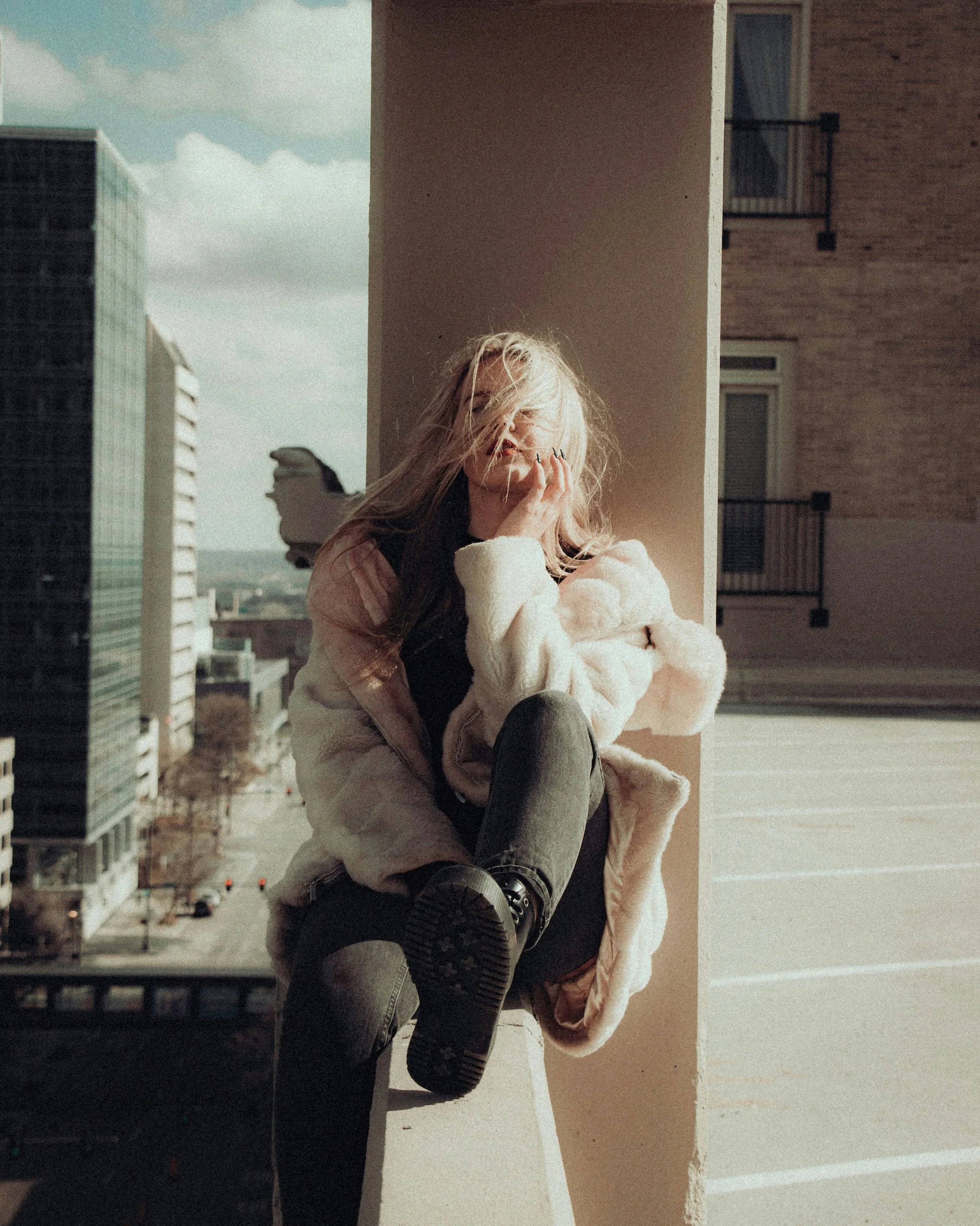A woman sitting on a balcony railing, with her head tilted back and hair blowing in the wind, overlooking a city street with modern buildings and a cloudy sky.