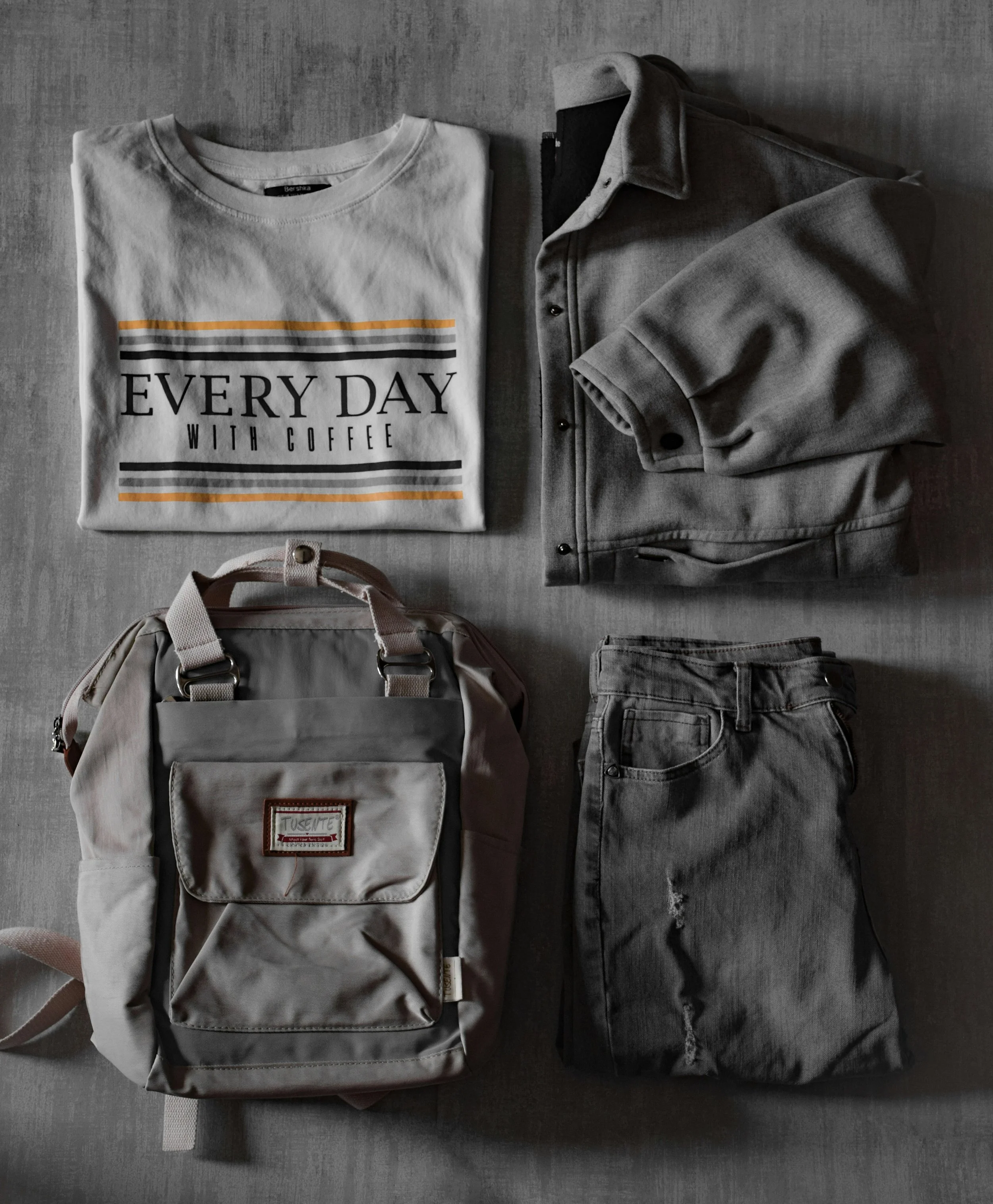 Flat lay of clothing items and accessories on a gray background including a white T-shirt with the text 'EVERY DAY WITH COFFEE', a gray denim jacket, a beige backpack, a pair of gray jeans, and a beige hat.