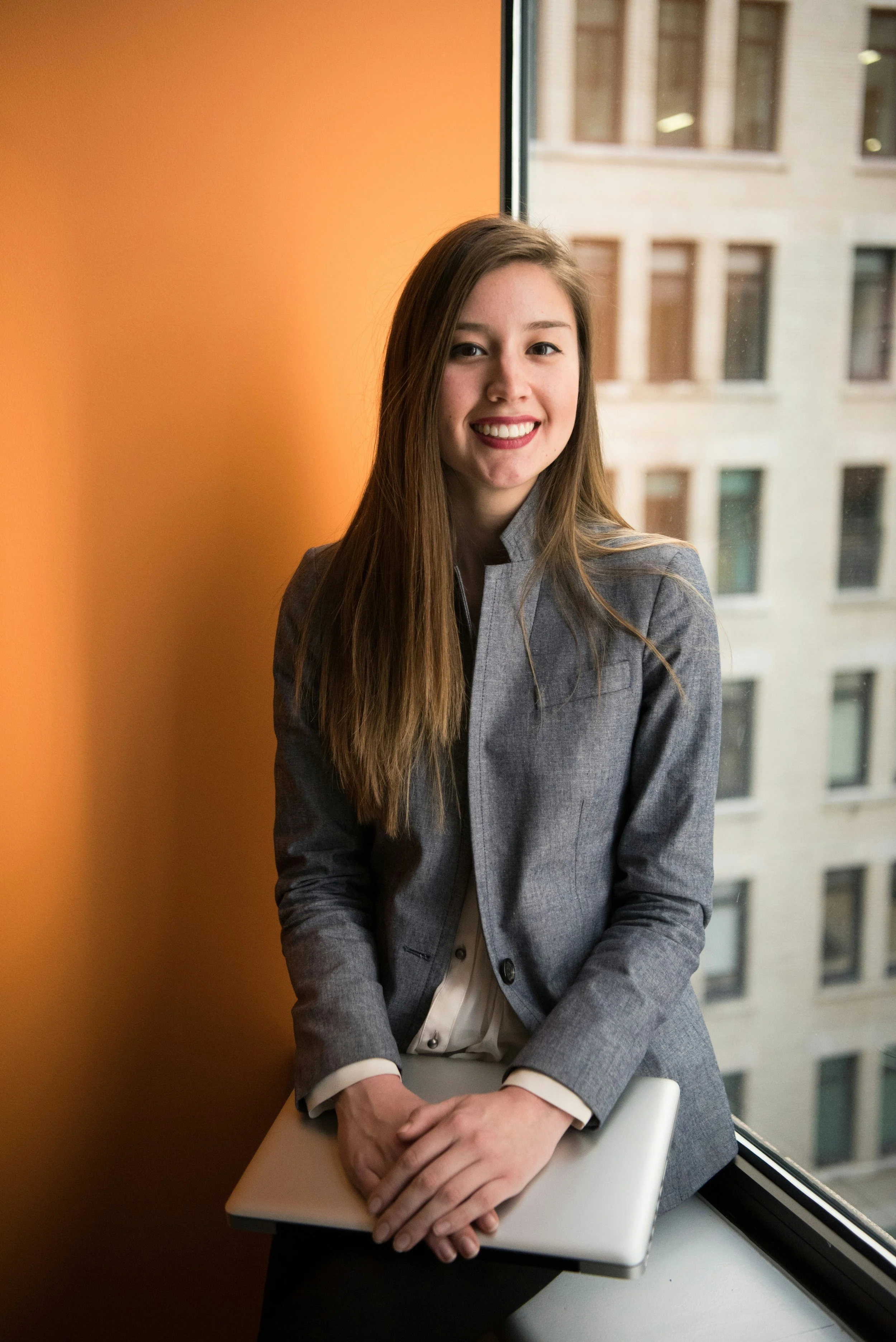 A young woman with long brown hair, wearing a gray blazer and cream-colored blouse, sitting by a window with a cityscape view. She is smiling, holding a closed laptop on her lap, and positioned next to an orange wall.